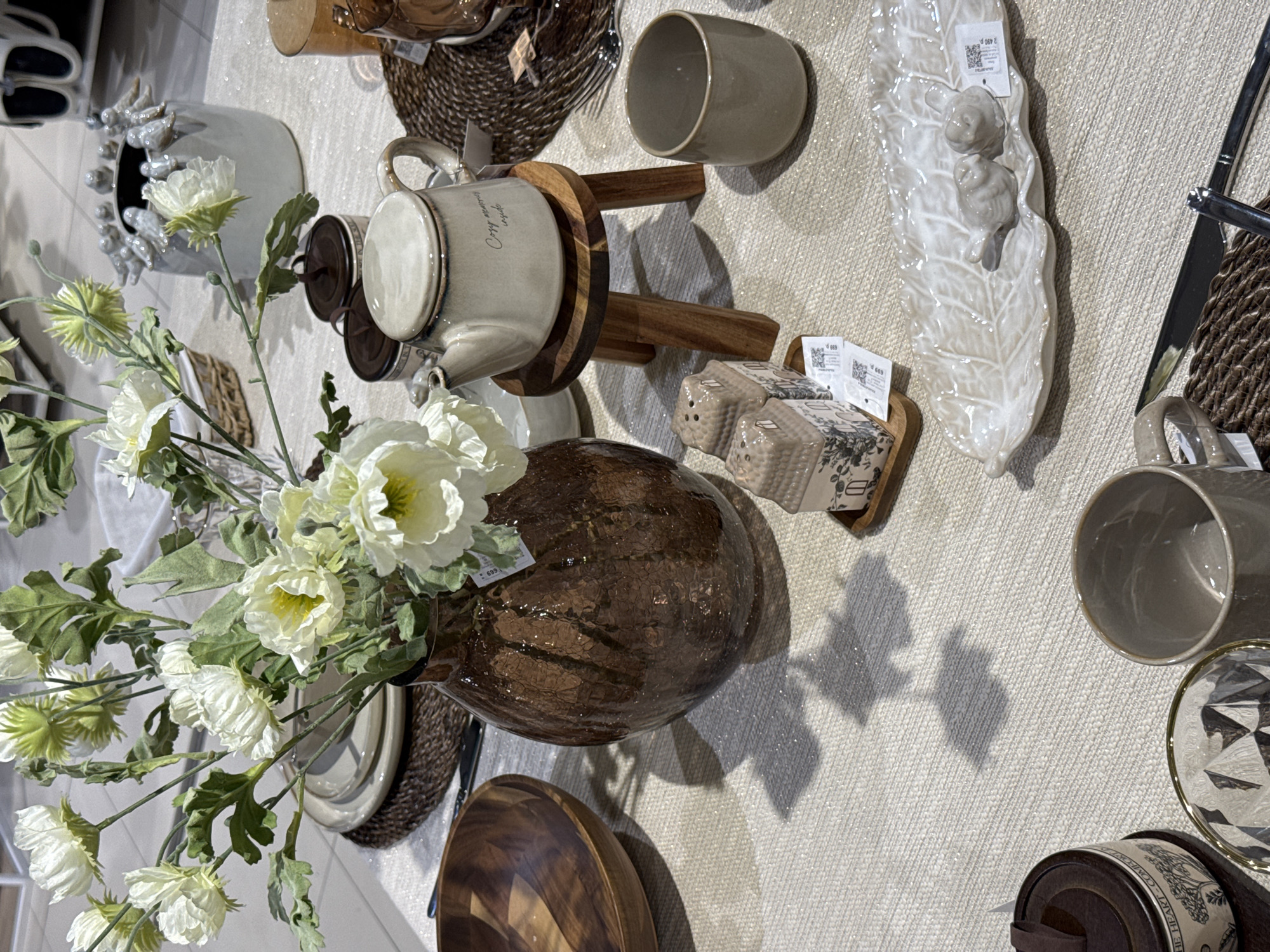 An overhead view captures a retail display table showcasing various home decor and dining items. The table is covered with a light beige, textured tablecloth exhibiting a subtle sparkle, upon which distinct shadows of objects and leaves are cast, indicating a light source from the upper left. In the bottom-left corner, a partial view of a cylindrical ceramic container with a dark brown lid is visible, inscribed with the partial text "THE HEART COMFORT." Adjacent to it, a faceted clear glass with a gold rim is positioned. A light taupe ceramic mug with a handle rests to the right of the faceted glass. A large, dark amber-colored glass vase with a textured, hammered finish serves as the central focal point on the left side, containing a bouquet of artificial white and light green flowers with green leaves. To the immediate left of the vase, a smoothly polished wooden bowl with visible grain is present. Directly in front of the vase, two miniature ceramic house-shaped shakers, decorated with dark grey botanical motifs, rest on a small wooden rectangular tray. A white price tag attached displays "699 ₽," indicating Russian Rubles. To the right of the shakers, a cream-colored, leaf-shaped ceramic dish features two small, light-colored ceramic bird figurines. Another white price tag, showing "1 699 ₽," is affixed to the dish. Below the leaf dish, a silver knife with a dark handle is placed on a round, dark brown woven placemat, which partially obscures a fork. In the center-right, a light grey ceramic teapot with a lid and handle, bearing the inscription "Cozy memories inside" on its side, is elevated on a small wooden three-legged stool. To the immediate right of the teapot, another light taupe ceramic mug is placed. Further right and slightly behind, two lidded dark brown ceramic containers, similar in style to the one in the bottom-left, are grouped together. In the upper-right background, a light grey ceramic vase is decorated with a ring of small bird figurines perched around its rim. Other out-of-focus retail display elements are visible in the distant background, suggesting a commercial environment such as a homeware store. The floor visible beyond the table is light-colored and tiled. No human subjects or interactions are present; all objects are static and arranged for display. Based on the visible Russian Ruble currency symbols (₽) on the price tags, this scene is located in the city of
