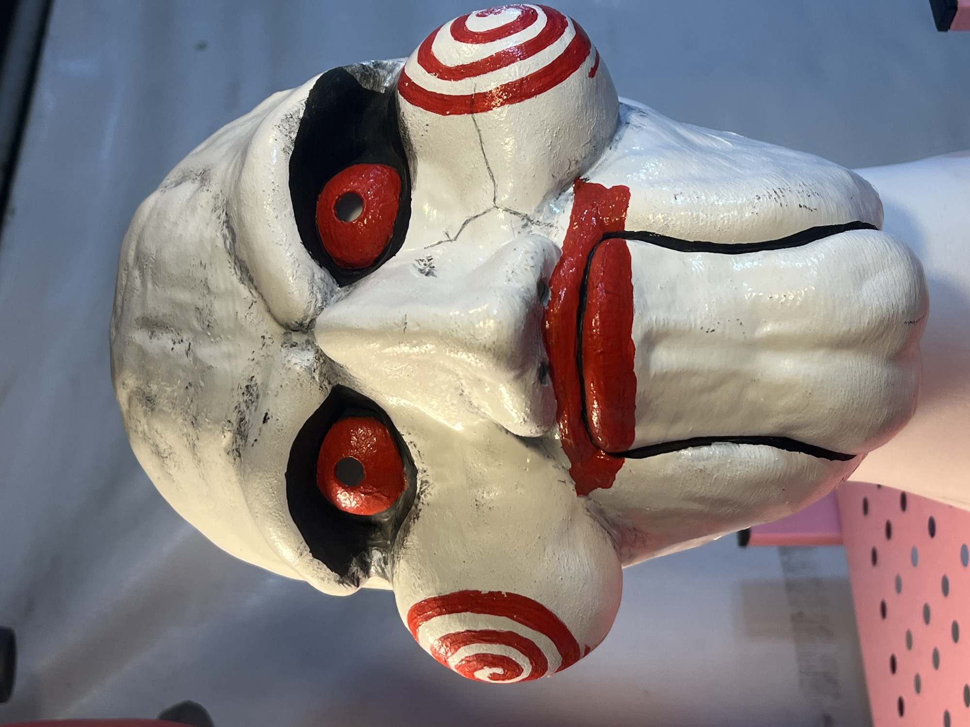 The image shows a close-up of a ventriloquist doll resembling the character Billy from the "Saw" horror franchise. The doll has a white, textured face with black eye sockets containing red, hollow eyes. Red spirals are painted on its cheeks and forehead. A crack is visible on the face, extending from near the nose towards one of the spirals. A thick red paint simulates blood running from the doll's mouth. The doll appears to be resting on a surface, with a pink object featuring black polka dots visible in the background. The location is unknown.