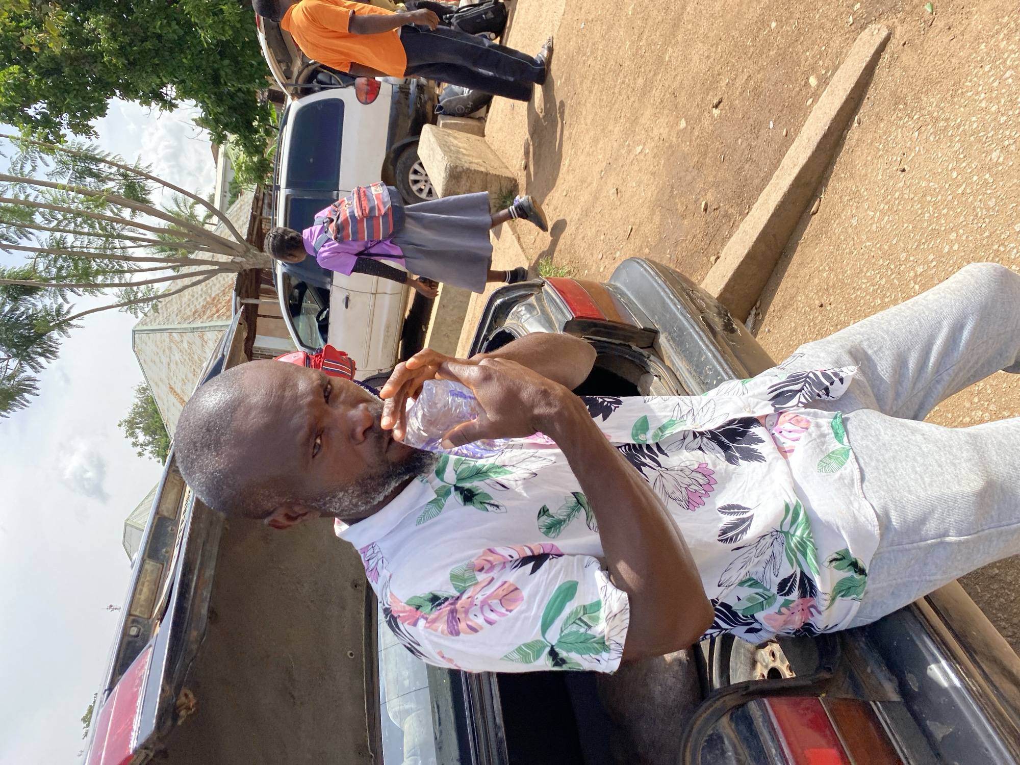 A man is shown from the chest up, leaning against the open trunk of a car. He is holding a clear plastic bottle to his mouth. He is wearing a white short-sleeved shirt with a floral pattern of pink, green, and black leaves and flowers. He has short graying hair and stubble. In the background, a young girl in a purple shirt and grey skirt with a backpack walks away from the camera. To the right, a man in an orange shirt stands near the open trunk of another vehicle. The scene appears to be outdoors in a dusty area, likely in Jos, Nigeria, with some buildings and trees visible in the background under a bright, partly cloudy sky.