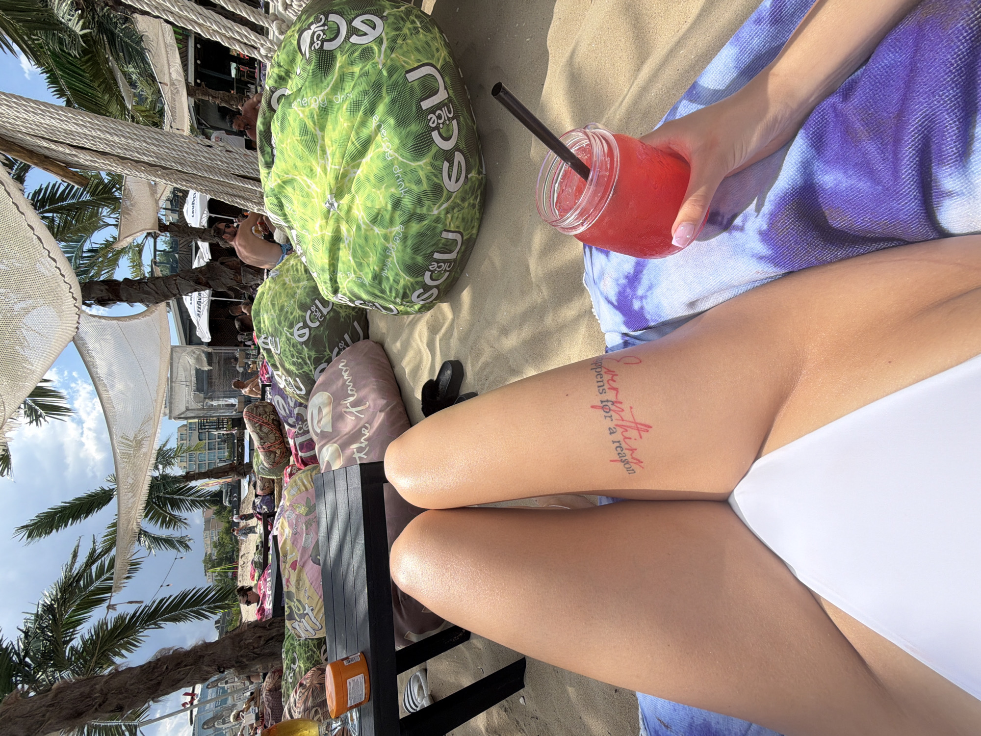 A first-person perspective shot shows a person lounging on a beach in Nesebar, Bulgaria, with their legs spread apart in the foreground, wearing a white bikini bottom. A tattoo on the person's left thigh reads "Everything happens for a reason" in pink cursive. To the right, a hand holds a red beverage in a mason jar with a black straw. The background reveals a crowded beach scene with large green beanbag chairs, palm trees, and tents. The sand is light brown, and the sky is bright blue with scattered clouds.