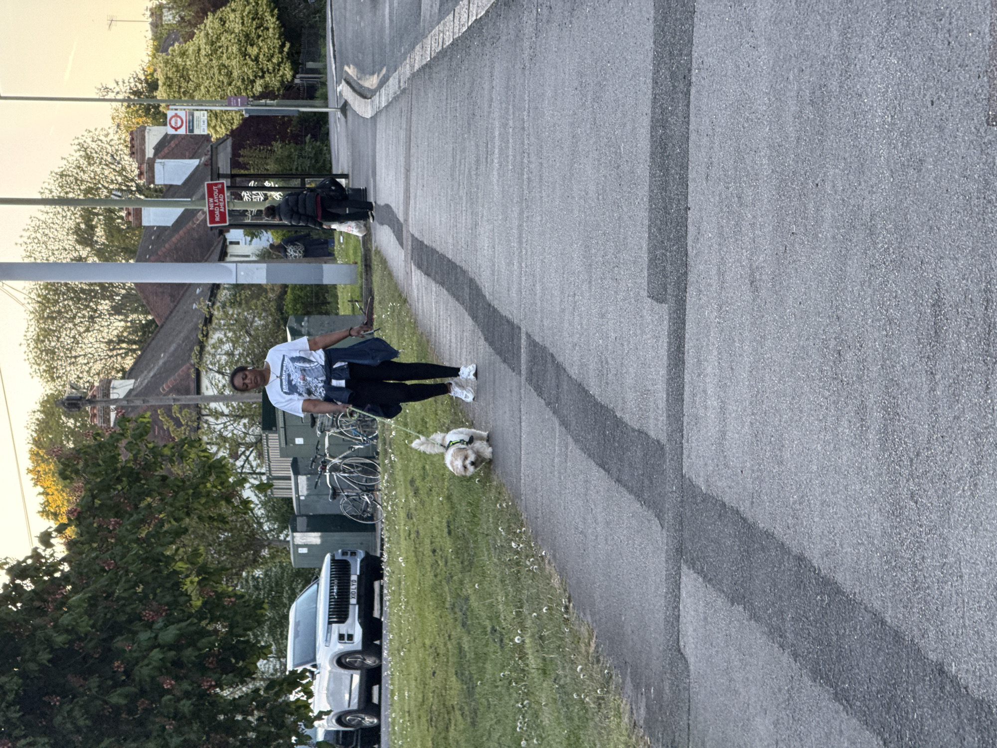 The image captures a sideways view of a person walking a dog on a paved path next to a road, seemingly in a suburban or residential area.
**Subjects:** The primary subjects are a woman and a small, fluffy white dog. The woman has dark skin, dark hair, and is wearing a white graphic t-shirt, dark leggings, and white sneakers. She holds a mobile phone in her left hand and a dog leash in her right, connected to the dog. The small dog, possibly a Shih Tzu or similar breed, wears a harness and walks obediently beside her. In the background, there are several parked bicycles leaning against a structure, and further back, a white SUV is partially visible. Houses with tiled roofs and lush green trees and bushes line the background. A bus stop shelter with glass panels is also visible.
**Activity:** The woman is engaged in the activity of walking her dog on a leash, proceeding along the paved pathway.
**Notable details:** The scene appears to be set during the late afternoon or early evening, characterized by a soft, golden light illuminating the tops of the distant trees and casting mild shadows. The weather seems clear and pleasant. The location cues strongly suggest the United Kingdom, specifically London, with the presence of a distinctive red London Underground roundel sign mounted on a pole. Other infrastructure includes streetlights/poles and standard road markings. The SUV's license plate is "X10 LYD". The overall image is captured with a sideways orientation.
**Visible text:**
* A red rectangular sign reads: "ROAD WORK AHEAD".
* A London Underground roundel symbol is visible, though specific station names are not legible.
* The license plate on the white SUV reads: "X10 LYD".