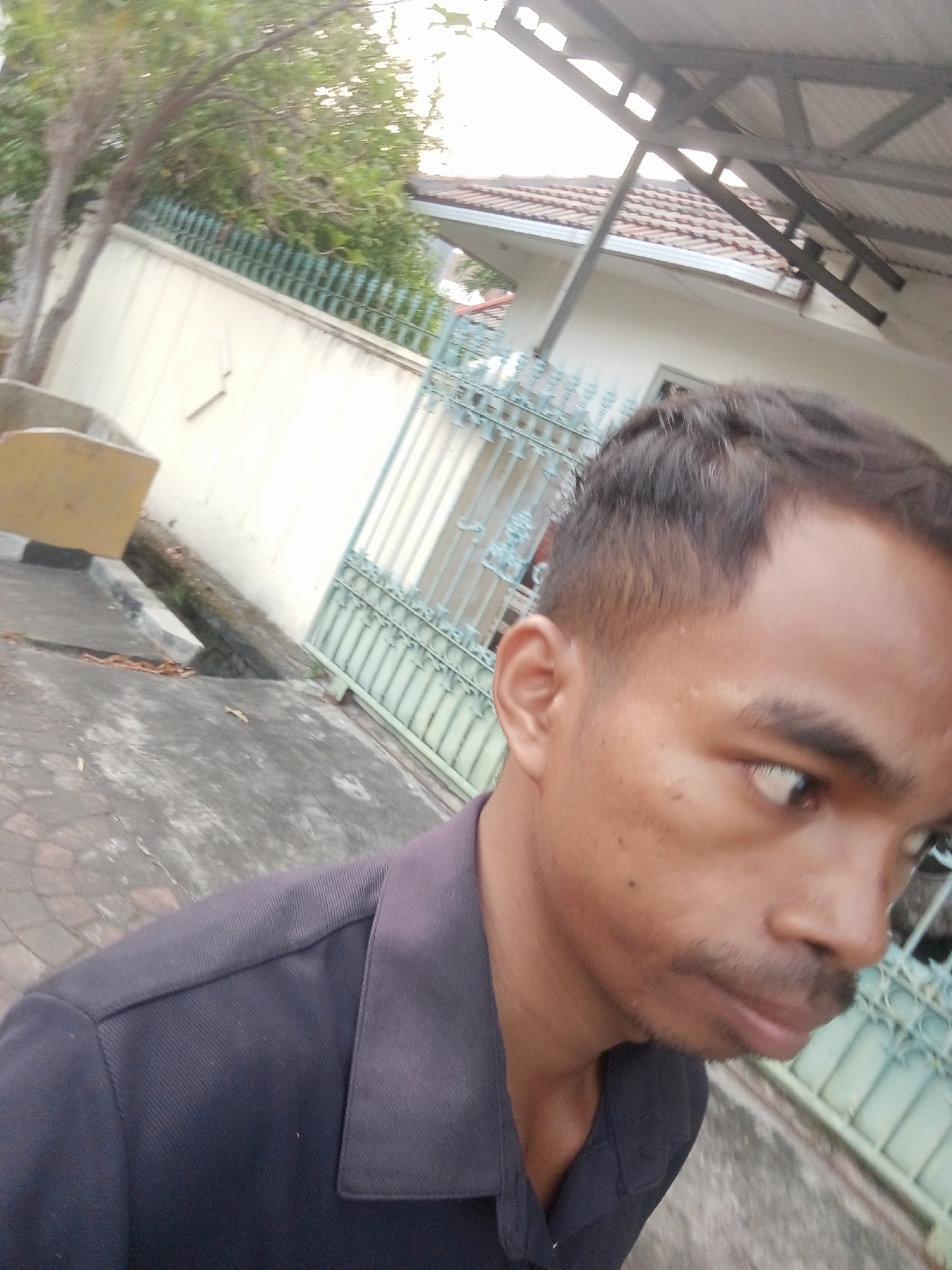 In Surabaya, Indonesia, a man is captured in a close-up, selfie-style shot outdoors. He is wearing a dark collared shirt. The background reveals elements of a residential setting, including a wall, a fence, trees, and a roof. The pavement in front of him is made of interlocking bricks.
