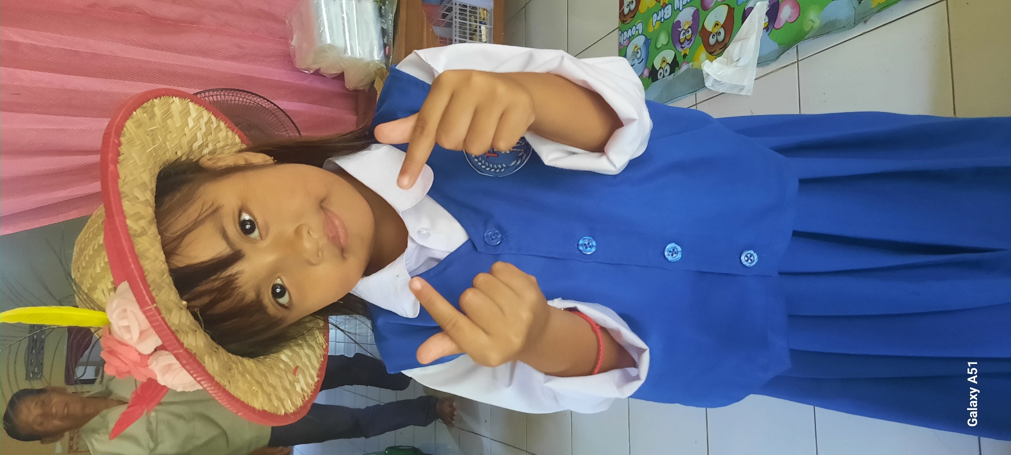 A young girl, likely of Southeast Asian descent, stands facing the camera, wearing a straw hat adorned with pink and red flowers and a yellow feather. She is dressed in a white collared shirt and a blue vest, which is buttoned up, over a blue pleated skirt. Her hands are raised, forming finger hearts. In the background, an elderly man in a light-colored shirt is partially visible. The setting appears to be indoors, with a pink curtain behind the girl and a tiled floor. A white plastic fan is visible to the right of the girl, and to her right, a table holds a stack of clear plastic-wrapped items and a colorful mat with cartoon owl characters. A red string bracelet is visible on the girl's left wrist. The scene is likely within a residential or educational setting in Ploso, Indonesia. The image is captured with a Samsung Galaxy A51 phone.