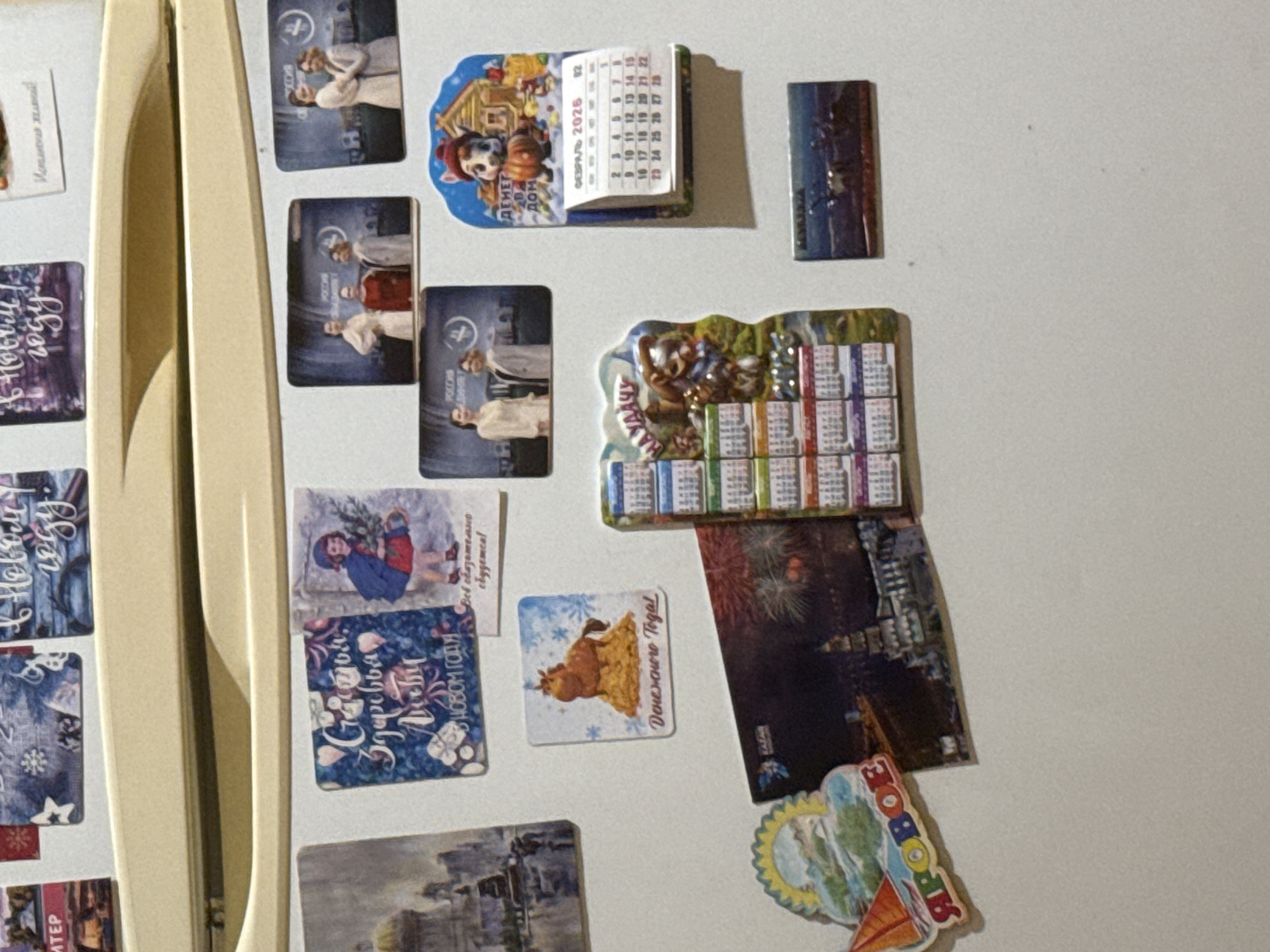 The image shows a collection of magnets on a refrigerator door. The magnets are varied, including holiday greetings, calendars, and scenic views. One magnet depicts a festive scene with a horse and snow, with Russian text that translates to "Happy New Year." Another magnet features a calendar for February 2026 with a cartoon character. A magnet with fireworks and a cityscape is also visible. There are no people visible in the image. The overall scene is domestic and appears to be in a home setting. There is no information to suggest the location is Kholmsk, Russia.