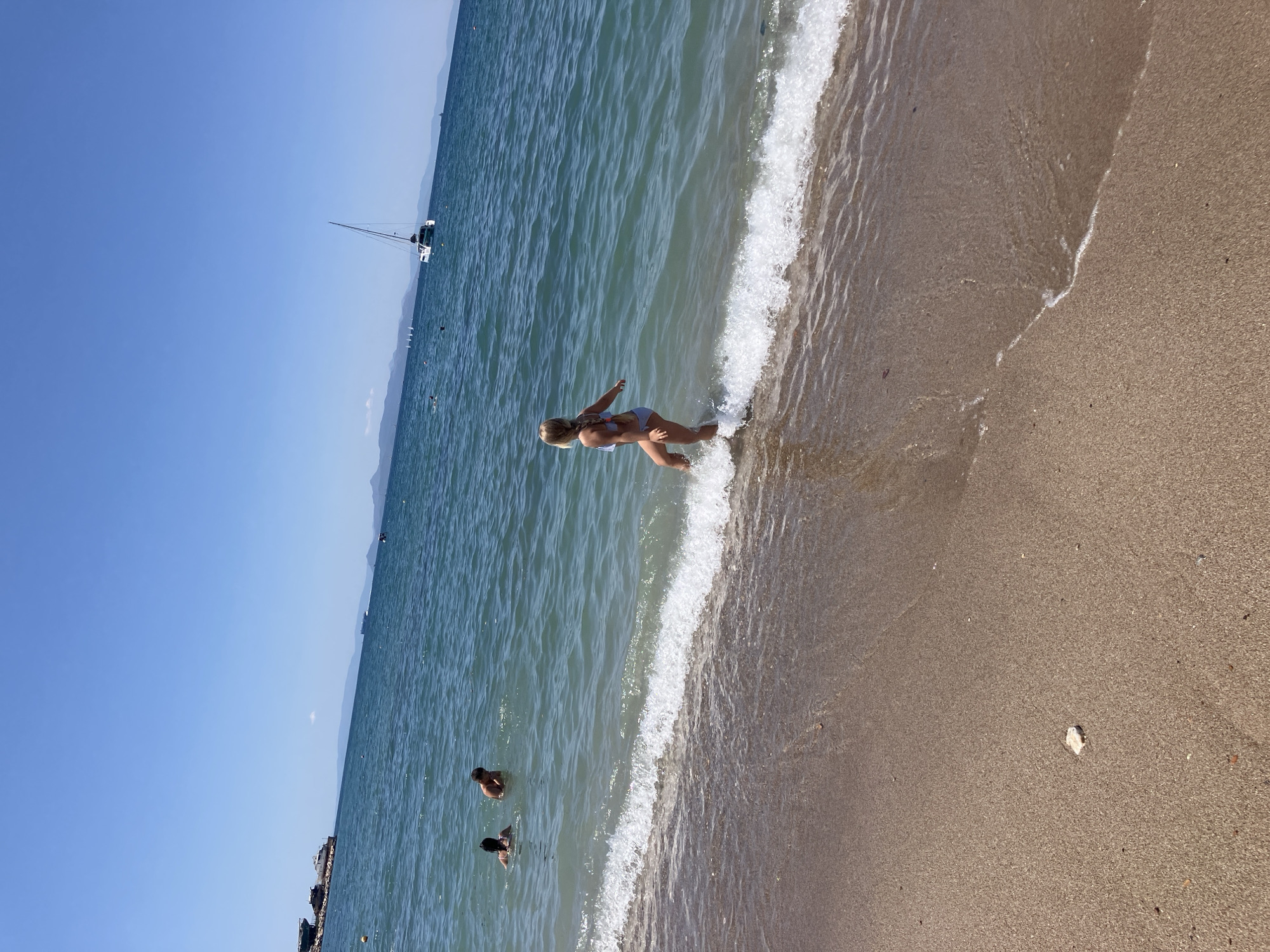 A photograph taken at Paleo Faliro, Greece, captures a beach scene on a clear, sunny day. The image is oriented vertically, with the sea on the left and the sandy beach on the right. The water is a vibrant blue-green, with gentle waves lapping the shore, creating white foam. A person in a bikini is visible standing in the shallow water, with their back to the camera, appearing to be mid-stride or movement. Further out in the sea, a few other individuals are swimming or wading. In the distance, a sailboat with a white hull and tall mast is anchored, and the hazy outlines of mountains form the horizon. The beach itself is a warm, sandy color, with small pebbles and shells scattered across it. The bright sunlight suggests it is midday or early afternoon. The overall mood is one of leisure and enjoyment of a summer day at the seaside. There is no visible text in the image.