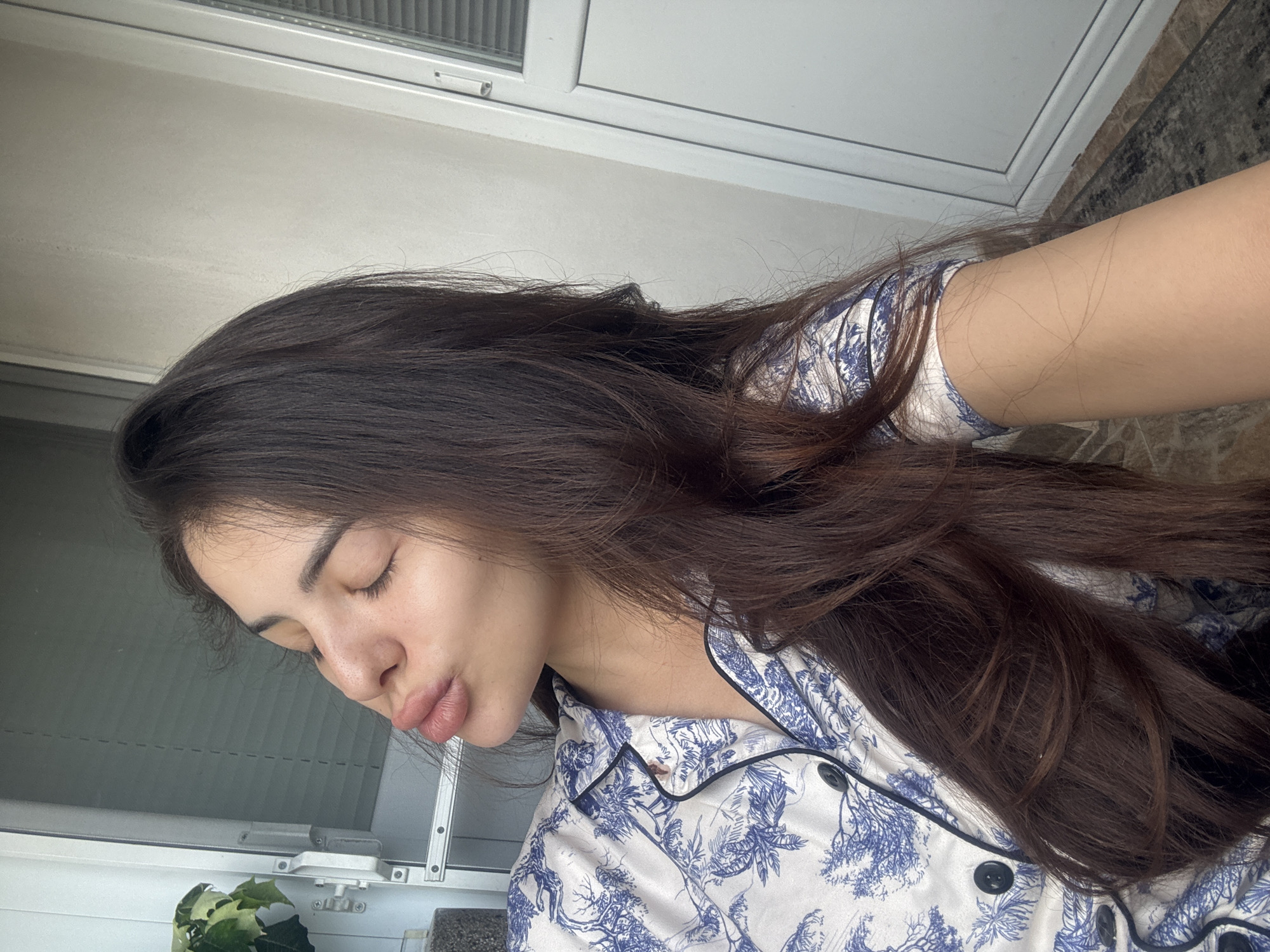 A young woman with long, dark brown hair is pictured from the chest up, leaning her head to the left with her eyes closed and lips puckered in a kiss pose. She is wearing a white pajama top with a blue toile pattern. The background shows a white wall and a section of a white door with vertical blinds. To the left of the woman, a plant is visible. The image appears to have been taken indoors near a window or balcony.