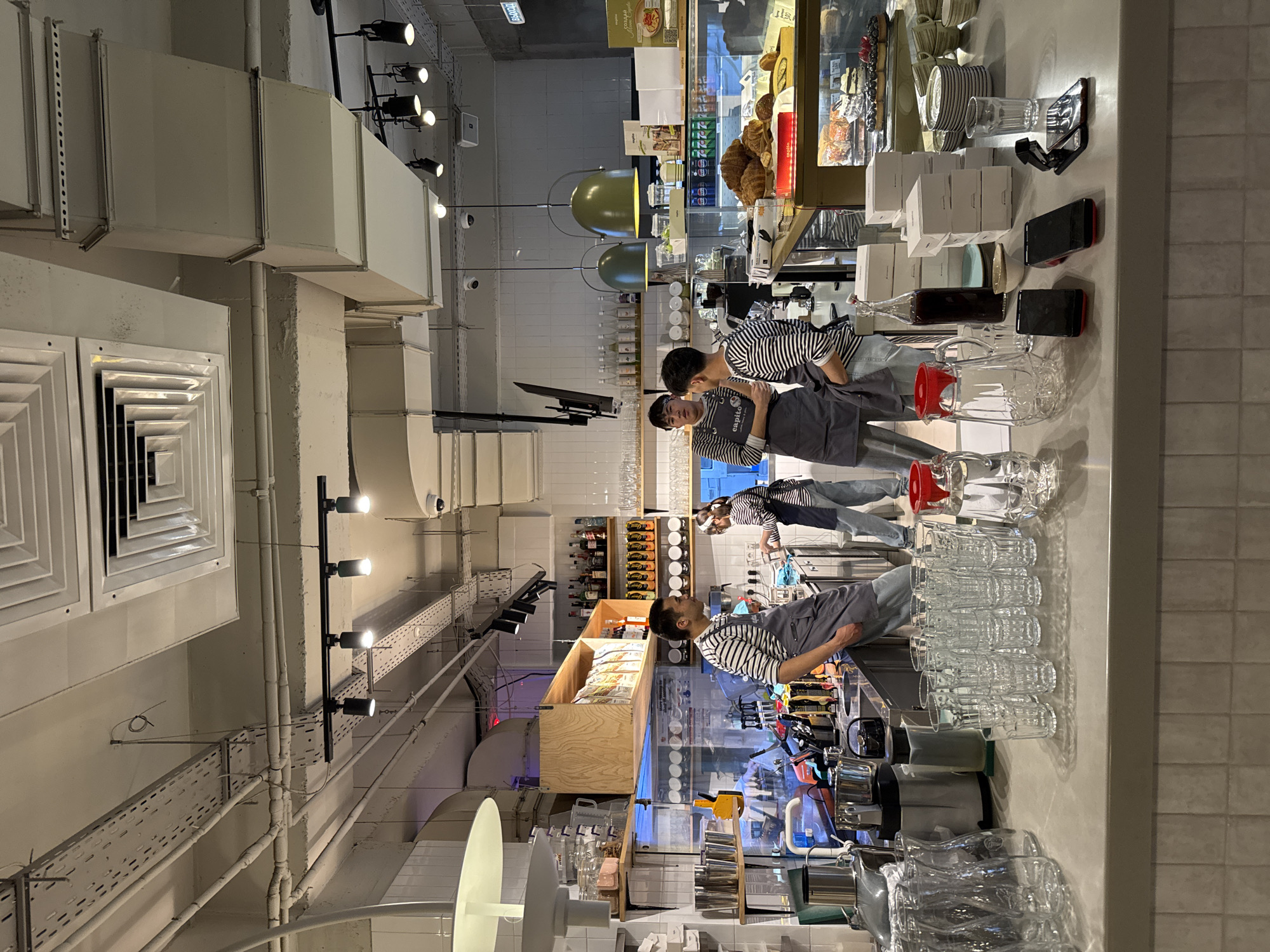 The image depicts the interior of a modern cafe or bakery in Bishkek City, Kyrgyzstan, characterized by an industrial-chic aesthetic. Three male staff members, dressed in striped shirts and dark aprons with "capito" visible on them, are positioned behind a long counter.
In the foreground, the counter is extensively equipped with numerous stacked clean glasses, pitchers with red lids, and what appear to be blenders or juicers. Several small white boxes and electronic devices, including smartphones, are also present. One staff member on the left is seen looking upwards or at a display, while another leans on the counter.
Behind the staff, a glass display case showcases an assortment of baked goods, including golden croissants and loaves of bread, alongside various bottled beverages. The background features open shelving stocked with bottles and other products. The ceiling reveals exposed white HVAC ducts, track lighting with multiple spotlights, and two large, olive-green pendant lamps that contribute to the contemporary industrial design. The walls are partially covered with white tiles.
The artificial lighting is bright, indicating an active operational environment. The staff appear focused on their tasks, suggesting a regular working scene. Visible text includes "capito" on the aprons and a poster within the display case.