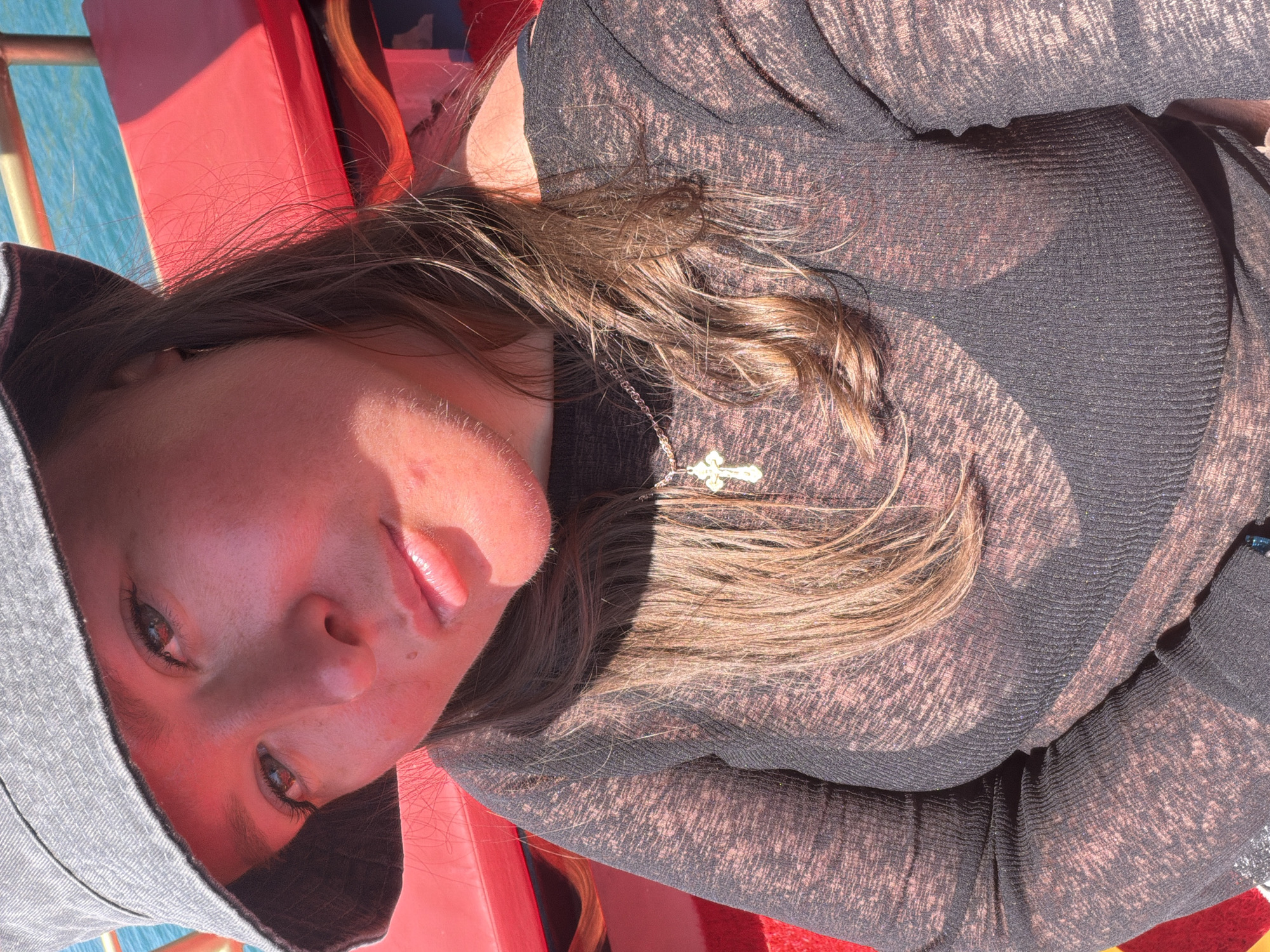 The image displays a close-up portrait of a young woman taken outdoors, rotated 90 degrees counter-clockwise, with her head positioned on the left side of the frame. She appears to be on a boat or near a body of water in Alanya, Turkey, under bright, direct sunlight.
The woman has long, medium-brown hair that flows over her shoulders and chest. She is wearing a dark, possibly sheer or textured, long-sleeved top, and a grey hooded garment with the hood pulled up, framing her face. Around her neck, a silver chain with a small, intricate cross pendant is visible. Her face is softly illuminated by the sun, showing a neutral expression with slightly parted lips. Her eyes are open and directed towards the camera.
In the background, bright red upholstered seating or cushions are prominent, suggesting the interior of a boat. A vibrant blue body of water is visible in the upper left portion of the image. The strong sunlight creates clear highlights on her skin and hair, imparting a warm, reddish tone to her face. The overall scene suggests a sunny daytime activity on the water. No text is visible in the image.