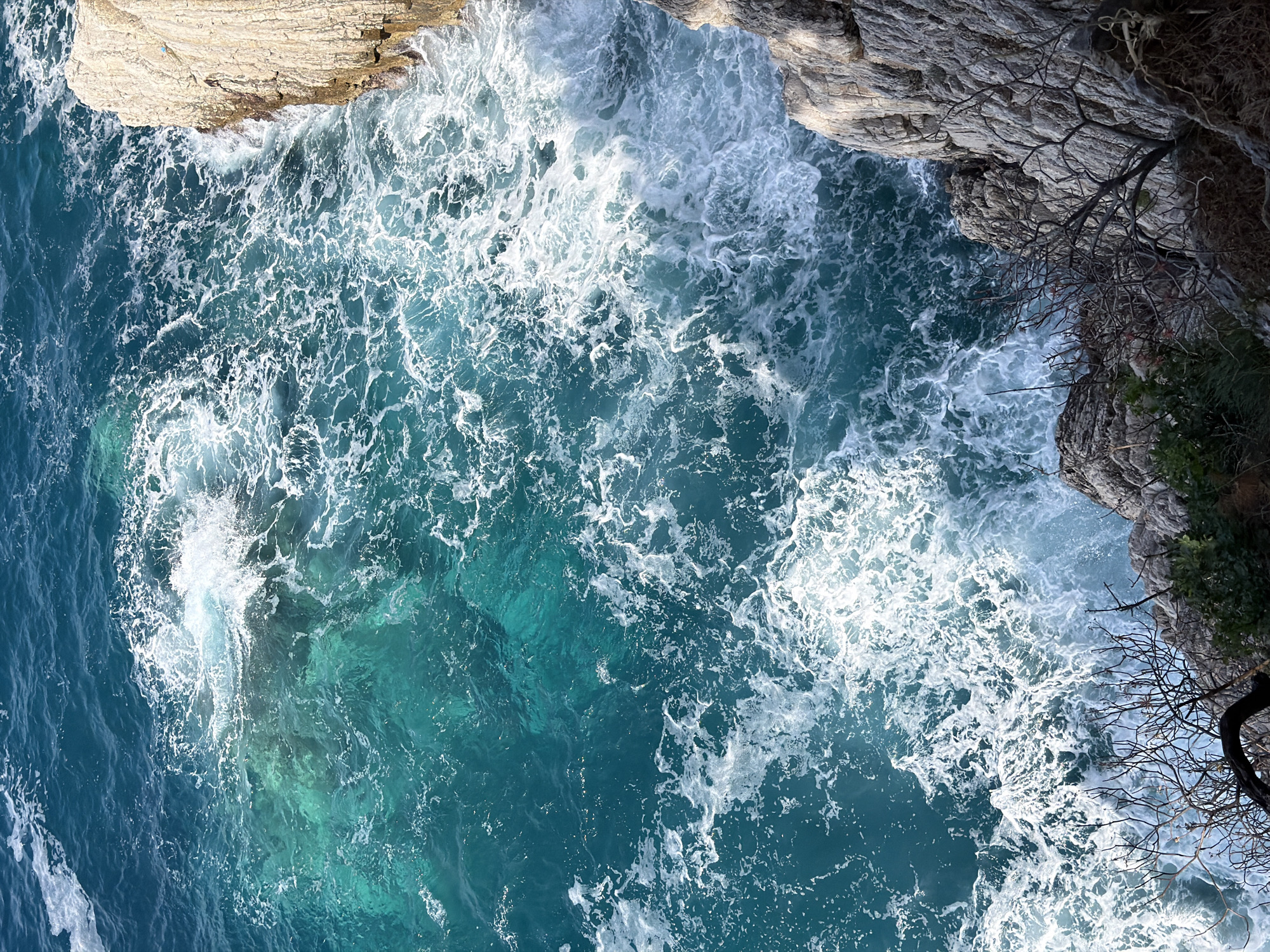 This image captures a high-angle, downward view of the turbulent sea crashing against rocky cliffs in Petrovac na Moru, Montenegro. The vibrant water displays a spectrum of blues and turquoises, ranging from deep azure in the deeper sections to clearer, lighter tones where light penetrates or sediment is visible.
White, frothy waves and extensive foam patterns dominate the water's surface, indicating significant wave action and turbulence, particularly near the coastline. The sea appears dynamic and powerful, with waves breaking and swirling.
Rugged rock formations frame the top and right edges of the image. These cliffs are composed of light-colored, textured stone, showing layers and crevices. Sparse vegetation, including some gnarled, bare tree branches and small green shrubs, clings tenaciously to the cliff faces, adding a touch of organic detail to the otherwise stark rocks. The lighting suggests bright daylight, illuminating the water's clarity and enhancing the textures of both the sea and the stone. There is no visible text in the image.