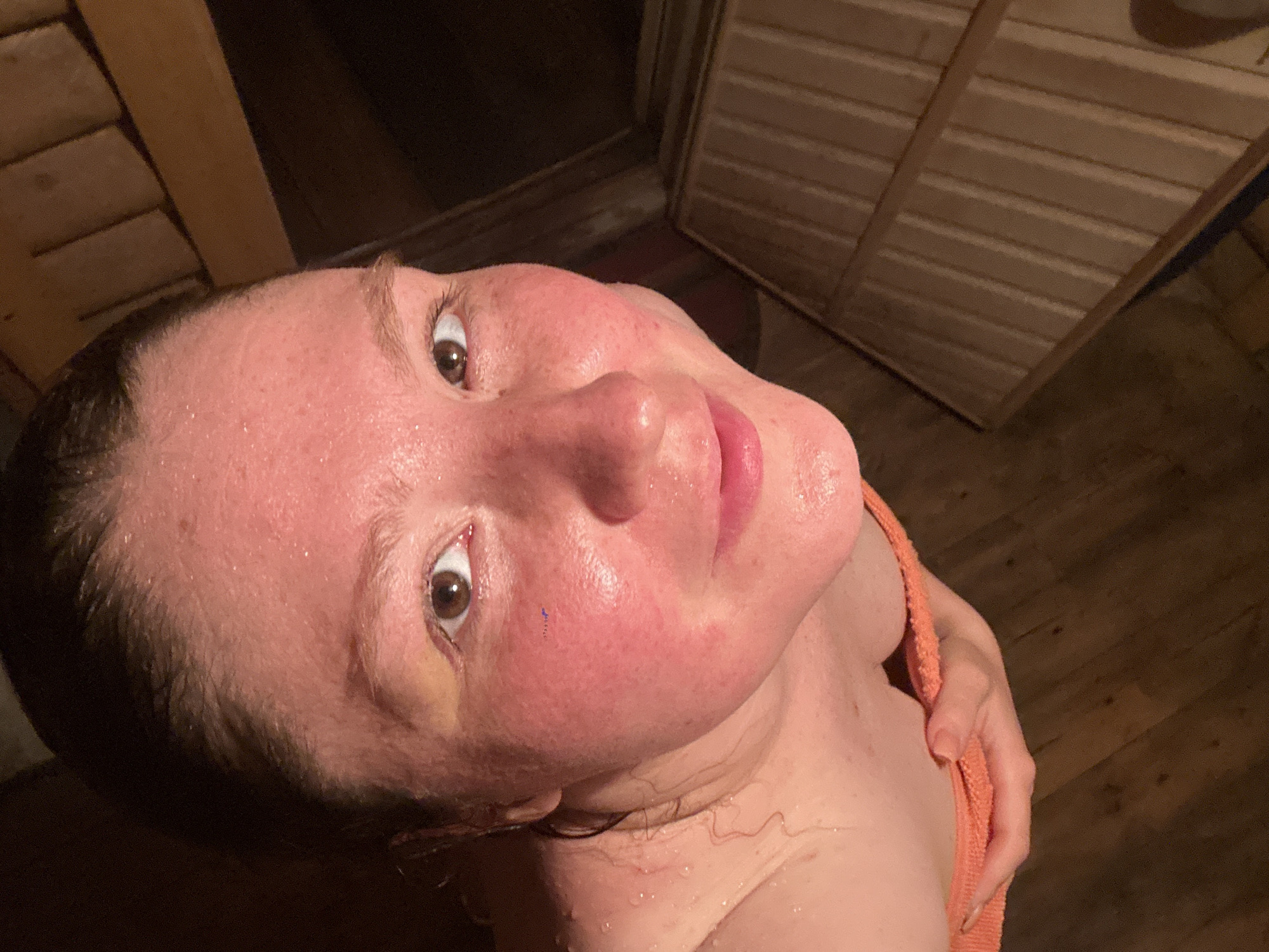 A close-up, high-angle shot shows a light-skinned, presumably Caucasian, woman in what appears to be a sauna or steam room. Her face is flushed and covered in beads of sweat, suggesting a high-heat environment. She has brown eyes. The woman is holding an orange towel against her shoulder. The background consists of wooden planks, typical of sauna interiors.
There is no way to determine whether the image was taken in the city of Nizhny Novgorod, Russia.