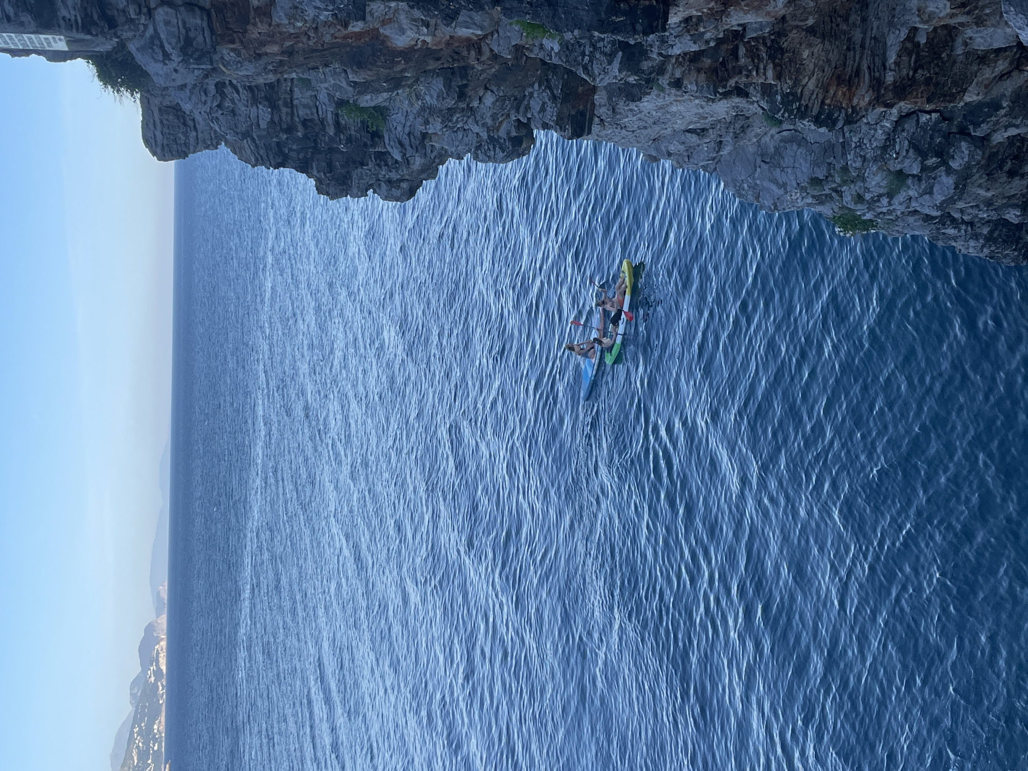A daytime aerial view oriented vertically captures two kayaks with people in the sea below an overhanging rocky cliff. The cliff occupies the top third of the frame. Two kayaks are located near the center, with three people visible on them. The water is dark blue and textured with small waves. The sky is a light blue at the left edge. On the horizon is the coastline. The scene is situated in Budva, Montenegro.