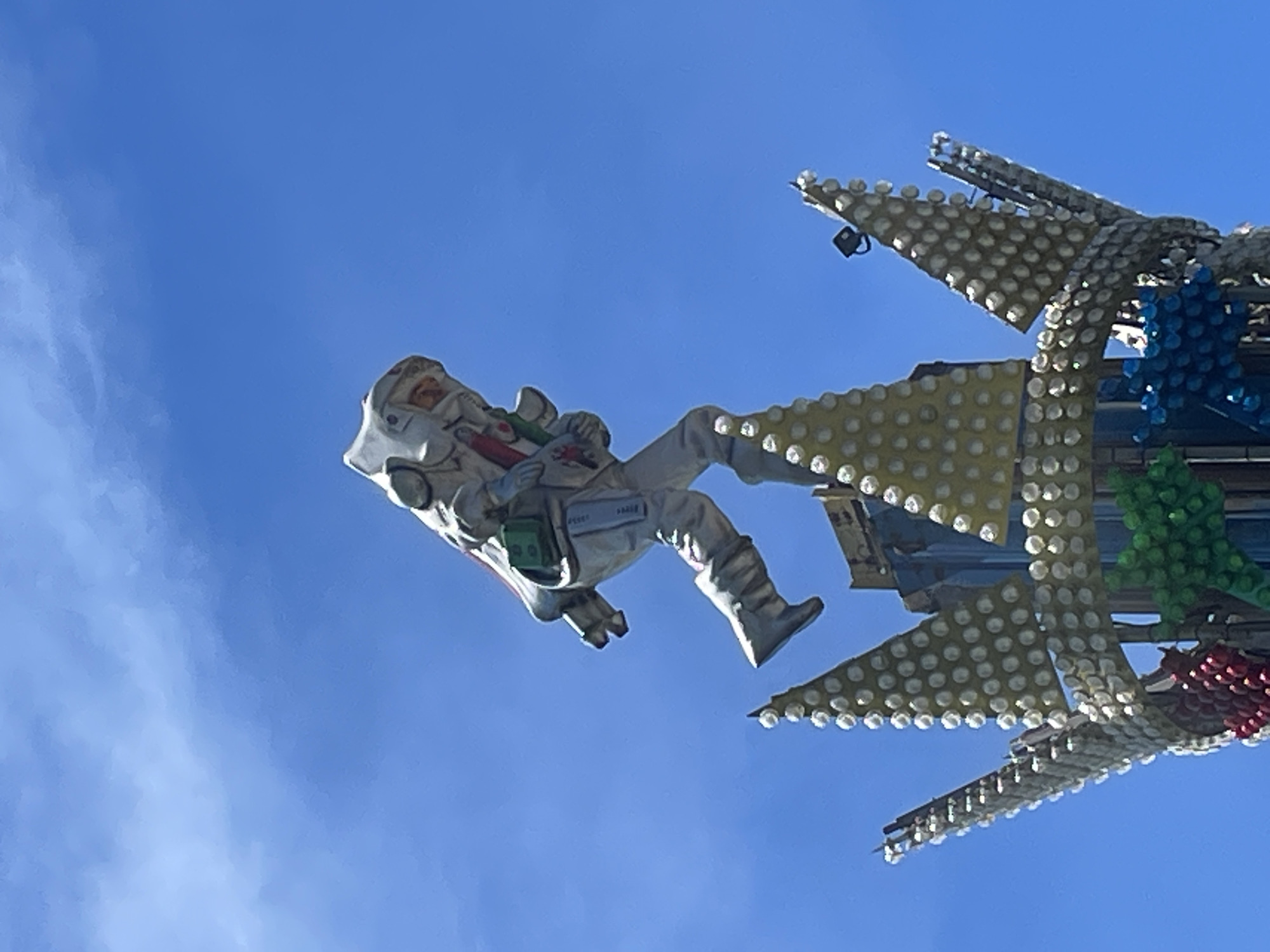 An outdoor, low-angle shot captures a decorative astronaut figure suspended against a bright, clear blue sky with scattered faint white clouds. The figure, dressed in a white spacesuit with a helmet, gloves, and boots, appears to be floating or in mid-flight. It holds two cylindrical objects, one red and one green, resembling small rockets or pyrotechnics. A green rectangular box is attached to its midsection, with faint text that appears to read "HARKER" or similar. Additional faded text is visible on the astronaut's right thigh, possibly "SPACE".
To the right of the astronaut, a large, complex structure is partially visible, adorned with numerous small, round lights, likely part of an amusement park ride or large-scale decoration. These lights are predominantly white/clear and arranged in clusters and rows on what appears to be a yellowish or golden paneling. Sections of this structure also feature blue and green ornamental elements, suggesting a vibrant, possibly space-themed or futuristic design. The angle of the shot, looking upwards, emphasizes the height and spectacle of the display.
The bright daylight suggests the image was taken during the day, though the extensive lighting on the structure implies it is designed to be illuminated at night. This scene is indicative of an amusement park or fairground attraction, consistent with a location like Sochi, Russia.