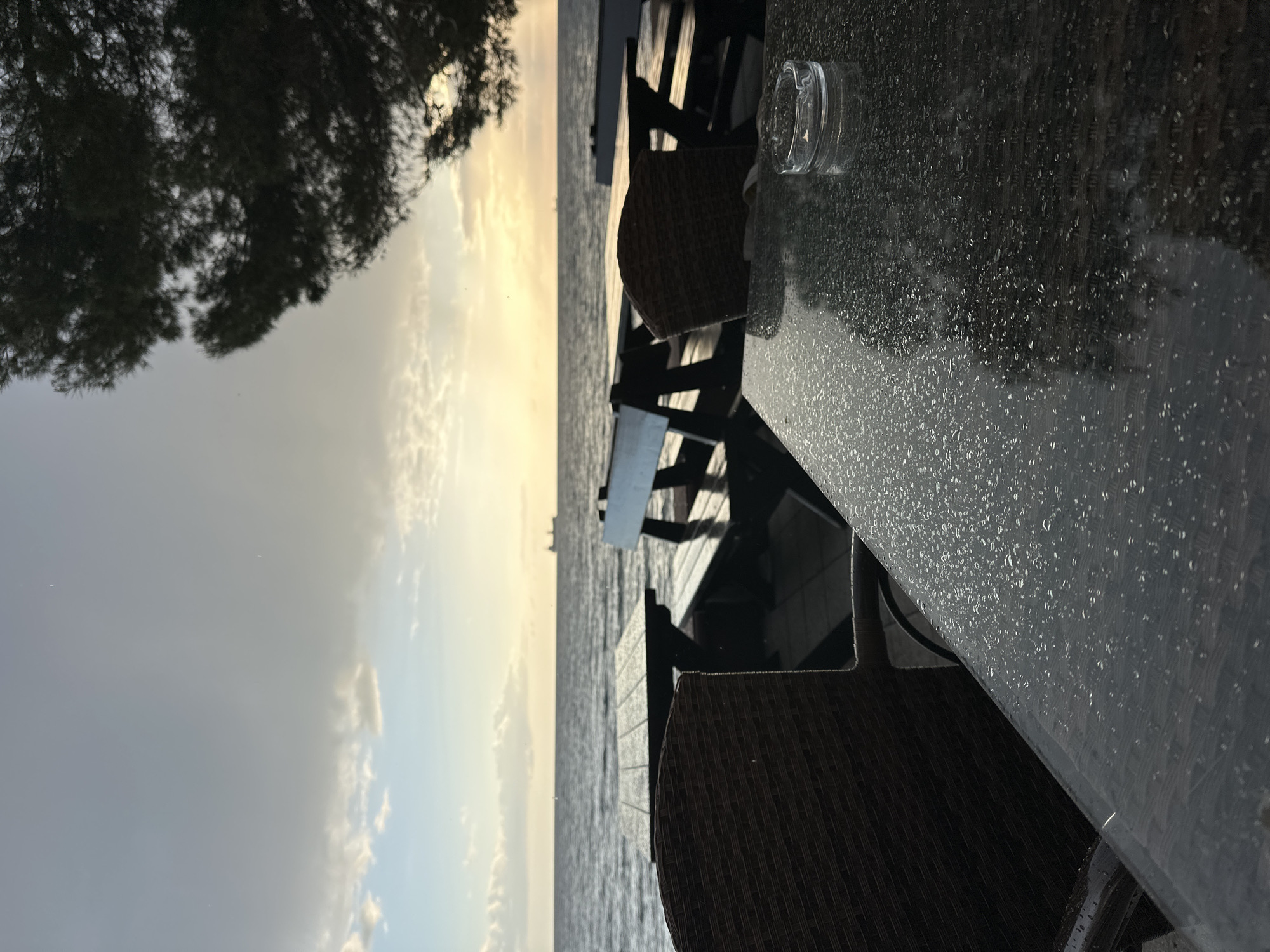 The image captures an outdoor seating area, likely a bar or restaurant patio, at either sunrise or sunset, overlooking a vast body of water. The perspective is from a table, looking out towards the horizon.
In the foreground, a dark, possibly stone or composite, tabletop is visible on the right, covered with numerous small water droplets, suggesting recent rain or heavy dew. A clear glass ashtray rests on the wet table. Dark, woven-style chairs are pulled up to the table. To the far left of the frame, the dark, silhouetted foliage of a tree partially obscures the sky.
The midground features more of these dark chairs and tables, extending towards the water's edge. The expansive body of water, likely the sea, stretches across the middle of the frame, appearing somewhat choppy with subtle reflections of the sky.
The background is dominated by the sky, transitioning from a bright, warm yellowish-orange glow on the left, indicative of the low sun, to softer, more subdued blues and grays with scattered clouds on the right. A faint, distant silhouette, possibly a ship or structure, can be seen on the horizon.
The overall atmosphere is serene and tranquil, with no people visible, suggesting a quiet moment. The wet surfaces hint at recent precipitation. Based on the provided context, the location is a bar in Montenegro, which aligns with the coastal setting depicted. There is no visible text in the image.