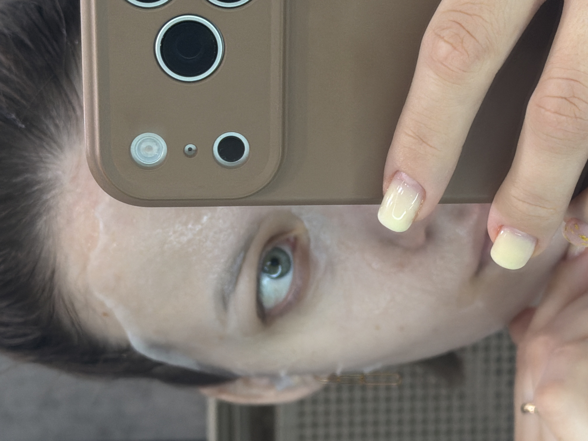 A person is taking a selfie with a smartphone in a brown case. The person's face is partially covered by a white mask. The person has light skin, dark hair, and greenish-blue eyes. The nails are painted a pale yellow color. The background is blurry, but there is a hint of a textured surface. The location is not discernible from the image, so it's impossible to confirm or deny its location in Reshetnikovo, Russia.