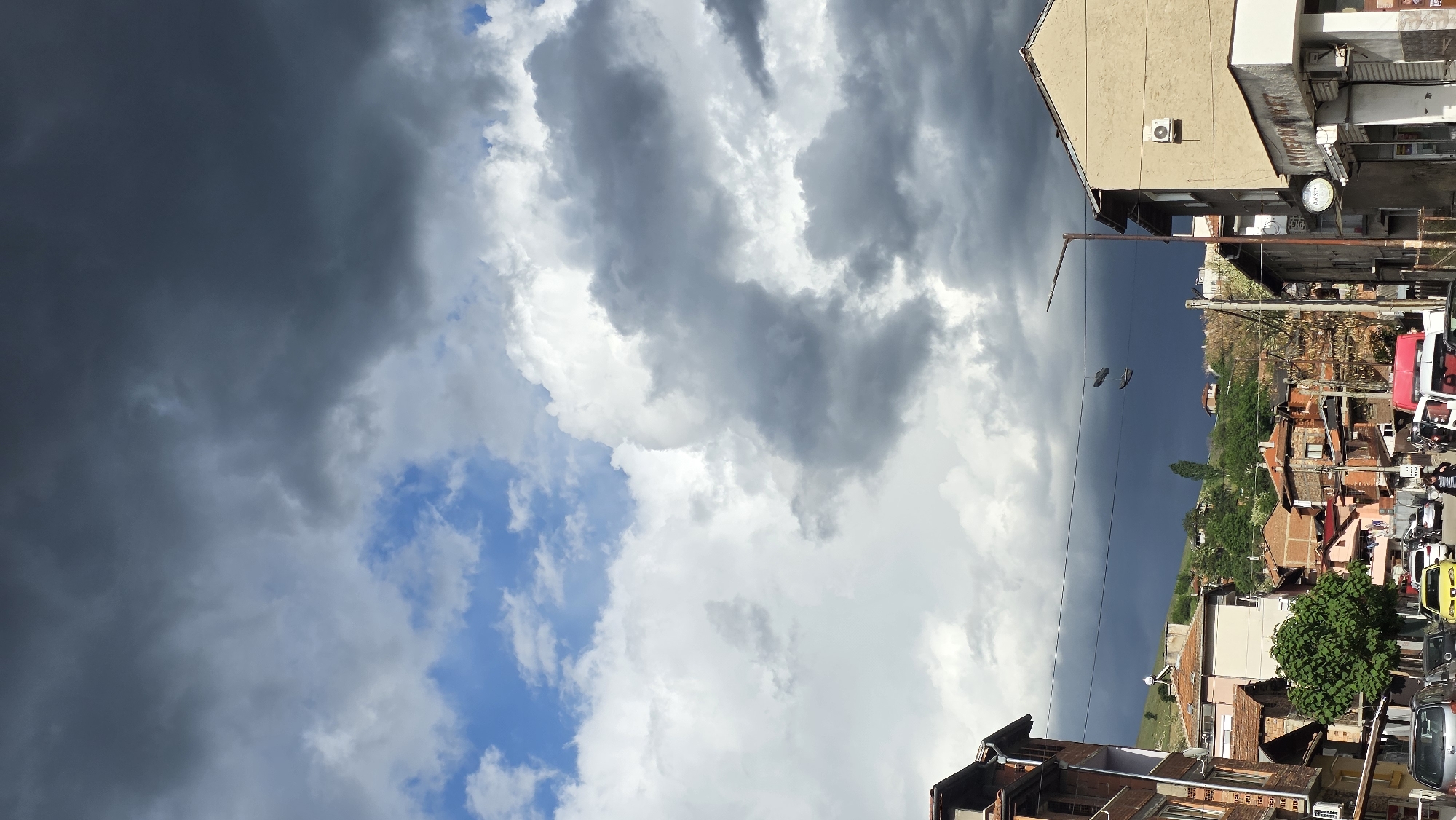 The image shows a partly cloudy sky above a street in Blagoevgrad, Bulgaria. The clouds range from dark gray to white. Buildings are visible along the street. Some rooftops are visible. Power lines cross the sky. Cars and trees are visible along the street.