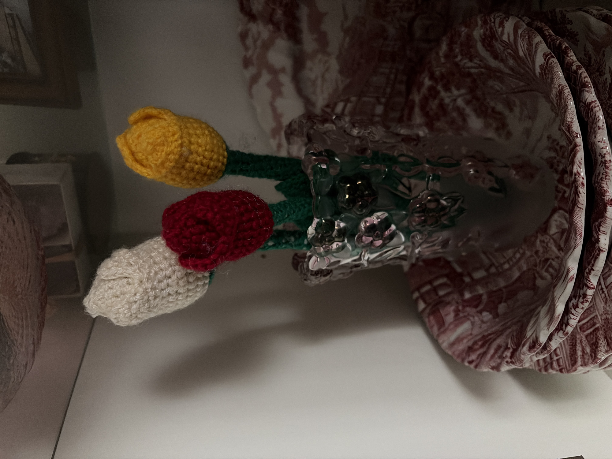A close-up, indoor, eye-level shot shows three crocheted tulips in a decorative glass vase. The tulips, one white, one red, and one yellow, are positioned in the center of the frame. The vase has a textured, clear glass appearance with a green stem visible inside. The vase sits on a stack of two plates, each with a red and white toile pattern featuring pastoral scenes. The background consists of a light-colored shelf and a muted, patterned fabric. The lighting is dim and atmospheric, casting shadows. The scene appears to be a decorative arrangement within a home or shop. There is no indication of the city of Astana, Kazakhstan.