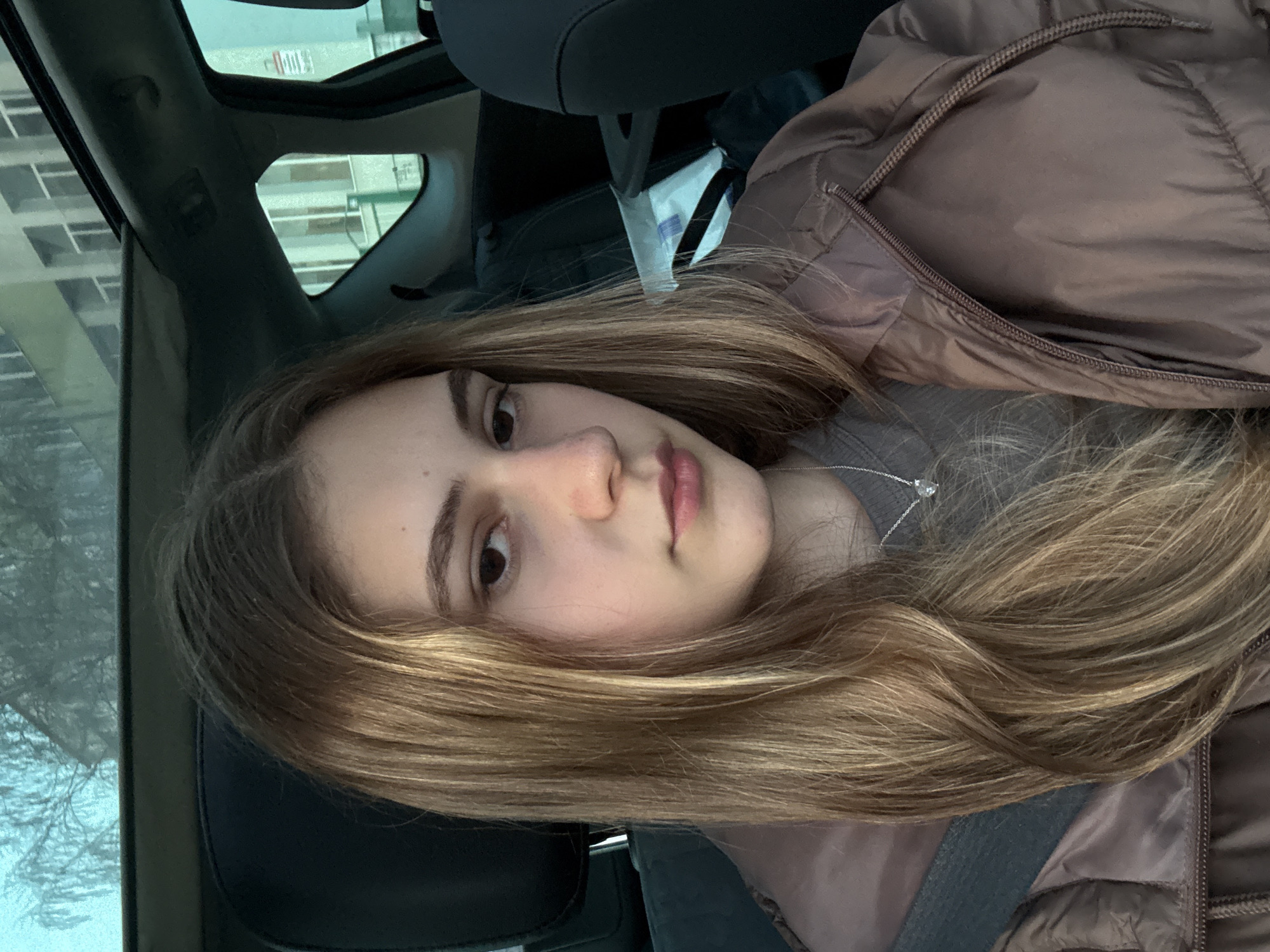 A young woman with long, light brown hair is sitting in the driver's seat of a car. She has a fair complexion, brown eyes, and a neutral expression. She is wearing a gray shirt, a brown jacket, and a silver necklace with a small pendant. The car's interior is visible, including the headrest, a seatbelt, and the car's roof. Outside the car window, a building with multiple floors and windows is visible. The exact location in Panagyurishte, Bulgaria, cannot be determined based on the provided image.