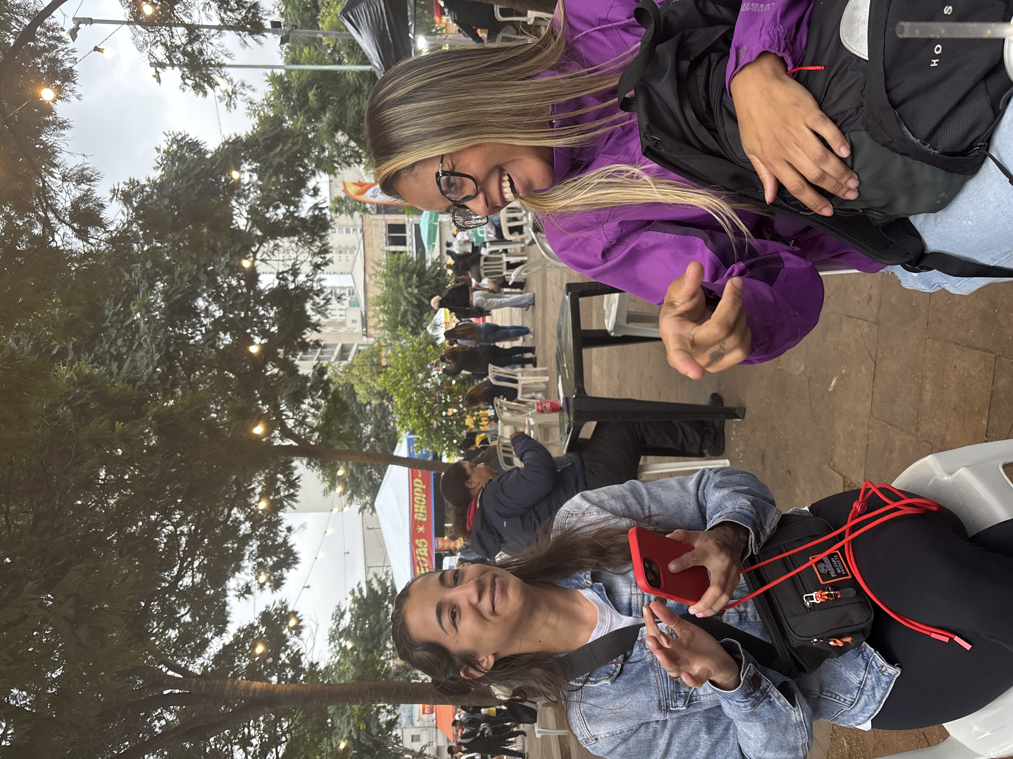 Two women are sitting at an outdoor cafe in São Paulo, Brazil. The woman on the left is wearing a blue jean jacket and black leggings. She has a red phone in her hand and a black bag with red cords across her lap. The woman on the right is wearing a purple jacket and has a black backpack. She is smiling and gesturing with her hand. The cafe is crowded with people and there are trees and lights in the background.