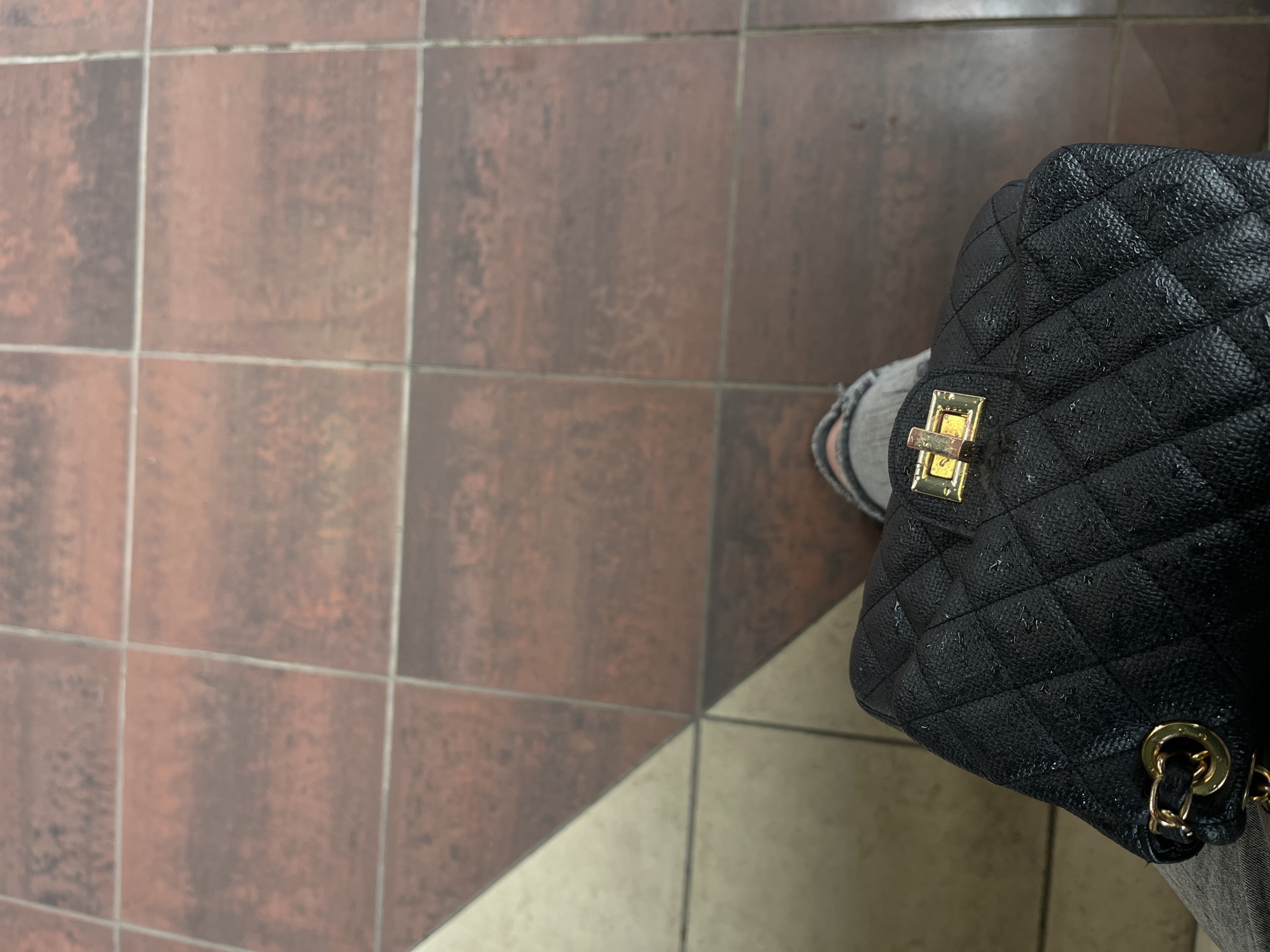 The image presents a top-down view of a black, quilted handbag resting on a tiled floor, positioned near the lower legs of an individual. The handbag features a pebbled texture, numerous small water droplets across its surface, a gold-toned rectangular clasp on its flap, and visible gold-colored O-rings at the strap's attachment points. The individual is wearing light grey trousers, appearing slightly distressed or worn near the hem, and dark footwear, with a glimpse of striped hosiery or lining visible above the shoe on the right. The floor is primarily composed of reddish-brown square tiles, exhibiting a varied, aged pattern and separated by light grey grout lines. In the lower-left quadrant, a distinct diagonal line separates the reddish-brown tiles from a section of lighter, possibly cream-colored, triangular tiles. This interior environment, characterized by its tile patterns and overhead lighting, is consistent with a public transit area, such as a metro station in Sofia, Bulgaria.