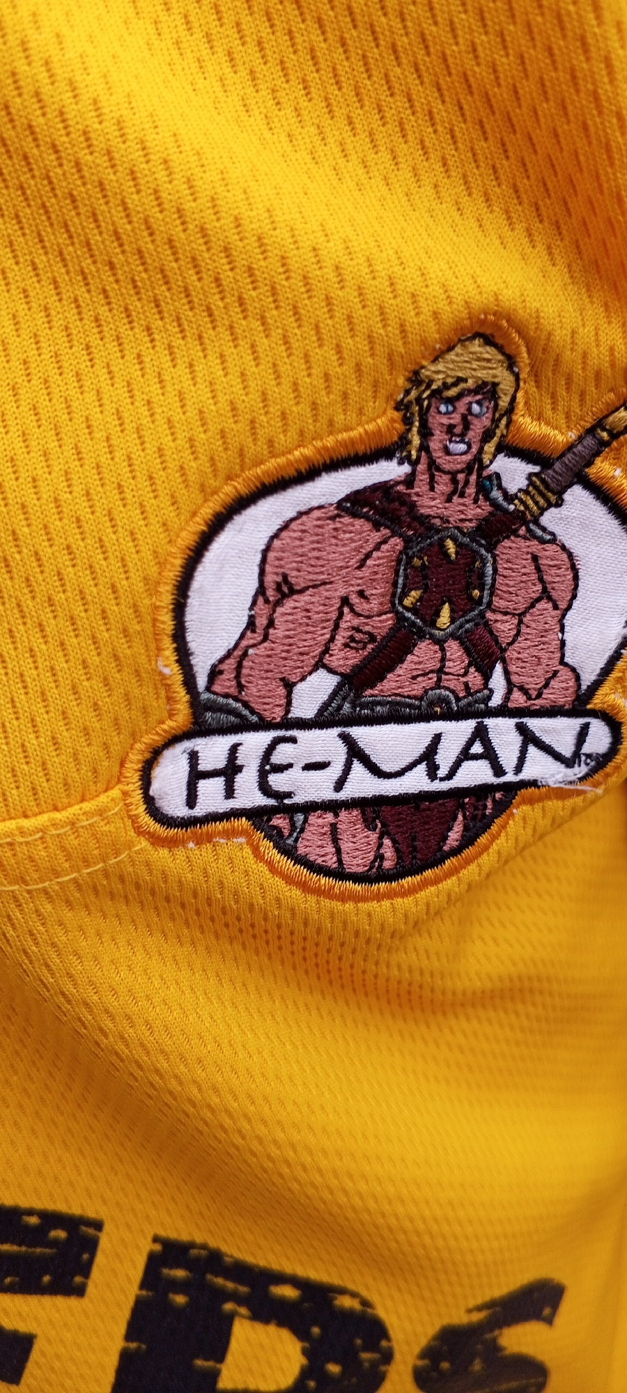 The image shows a close-up of a yellow, mesh-like fabric, likely part of a garment. Embroidered onto the fabric is a patch featuring the character "He-Man" from the Masters of the Universe franchise. The patch depicts He-Man from the chest up, with his muscular physique, blond hair, and signature armor and sword. Below the character, the text "HE-MAN" is embroidered in black lettering on a white background, outlined by a yellow border. The fabric shows a diamond-shaped mesh pattern and a horizontal seam is visible on the left side of the image, suggesting this is a piece of clothing. The image is cropped tightly on the embroidered patch and the surrounding fabric, with the background out of focus. There is no discernible indication of the city of Jalingo, Nigeria within the image.