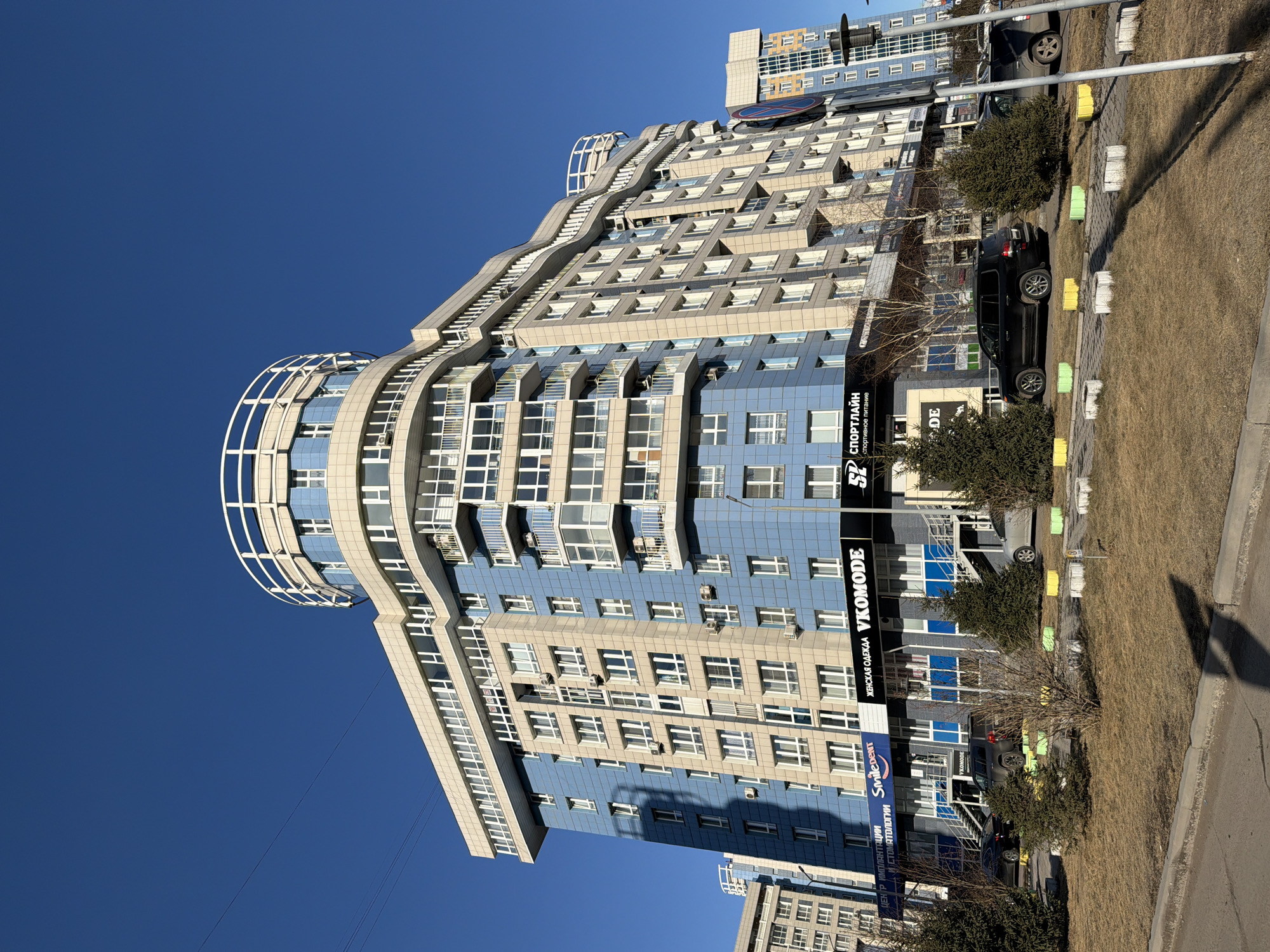 The photograph shows a multi-story building in Krasnoyarsk, Russia. The building's facade is primarily blue and beige, with many windows and balconies. A distinct architectural feature is a semi-circular structure at the top. Signage is visible on the building, including one that says "VKOMODE" and another with Russian text; another says "SPORTLAIN спортивное питание". There are two vehicles parked nearby, a black SUV and a light-colored sedan. The surrounding environment includes dry grass, trees, a sidewalk with yellow and white markers, and a clear blue sky. Another building can be seen in the background. No people are clearly visible in the image. The angle of the photograph appears to be from ground level, looking upwards.