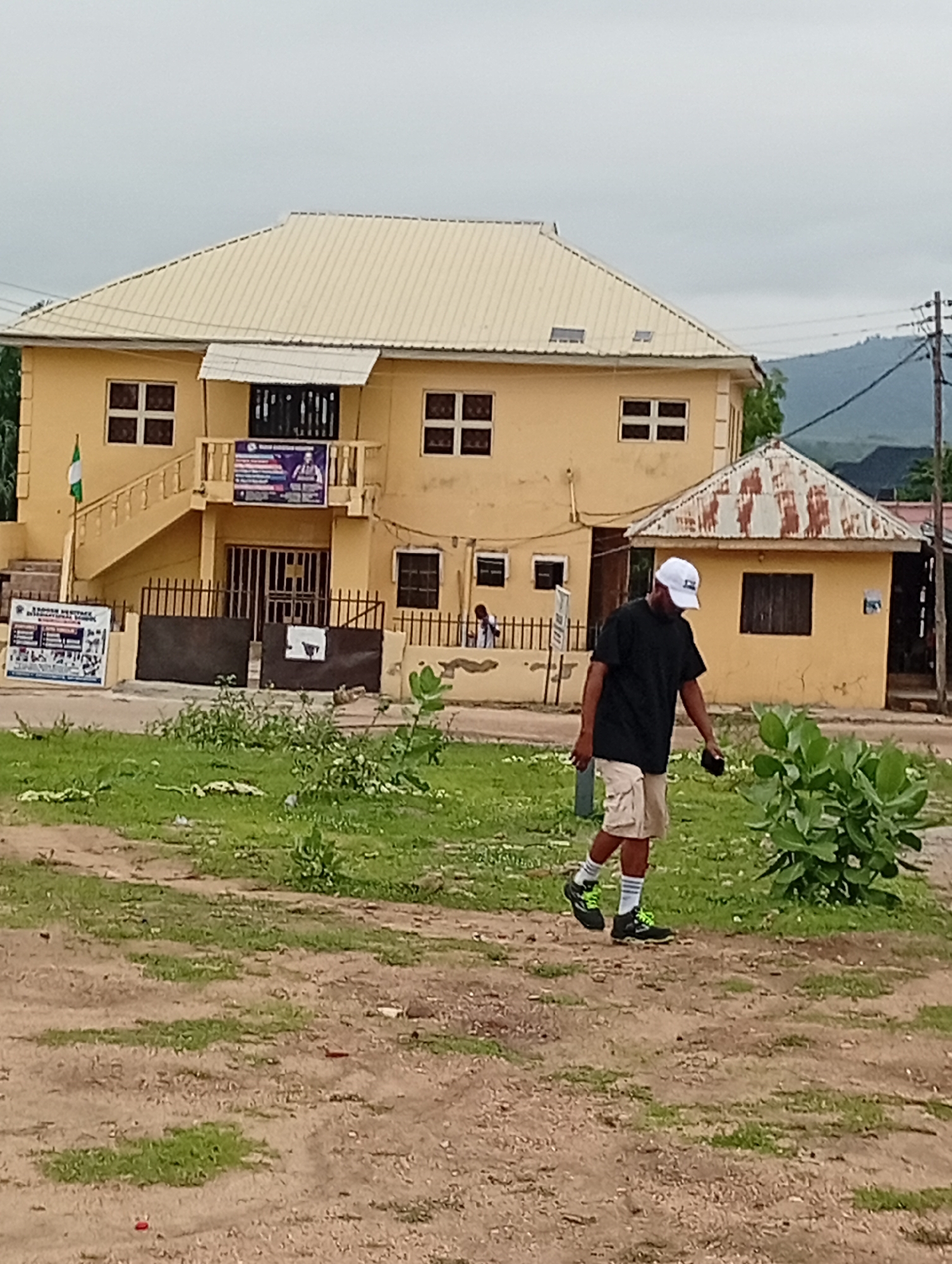 A medium shot depicts an individual walking on a dirt and grass-covered area in what appears to be a public space. The person, wearing a black t-shirt, khaki shorts, a white baseball cap, and black sneakers with green laces and white socks, appears to be looking at something in their hand. In the background is a two-story yellow building with a beige roof and a smaller, single-story building attached to it. A green Nigerian flag is visible near the left side of the main building. The building is surrounded by a metal gate. To the right of the main building, a mountain range is visible in the distance. The scene is likely located in Lokoja, Nigeria.