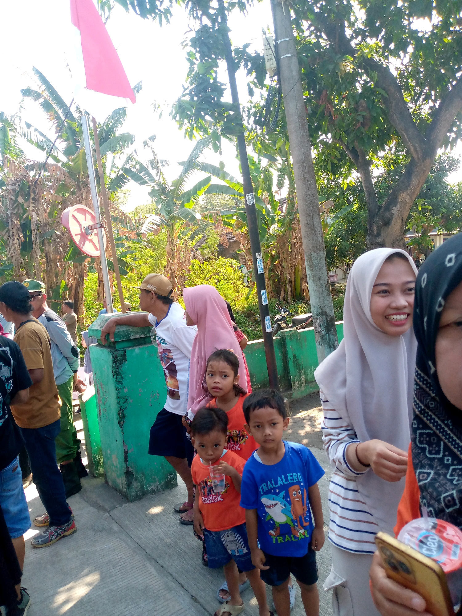 In the city of Sumberwono, Indonesia, a group of people are gathered outdoors on a sunny day. In the foreground, a young boy in an orange t-shirt and blue shorts drinks from a clear cup with a straw. To his left, another young boy wears a blue t-shirt with a shark graphic and the text "TRALALERO" and stands with his hands by his sides. Behind them, a young woman in a light gray hijab smiles broadly as she looks to her left, her right hand resting on her chest. Next to her, the right side of another woman's face is visible, wearing a black hijab with a patterned trim.
A group of adults stand further back. A man in a tan baseball cap and a white t-shirt leans on a green concrete structure. To his left, a woman in a pink hijab stands. Other adults are partially visible, including one in a tan t-shirt and blue jeans on the far left.
The environment consists of a paved walkway or road, with a weathered green concrete structure along the right side of the frame. In the background, lush green vegetation, including banana trees, and trees with dense foliage are visible under a bright, possibly overexposed sky. A utility pole with wires runs vertically along the right side of the image. A red and white flag is attached to a pole on the left side of the frame. The overall impression is a casual outdoor gathering, possibly a community event or celebration.