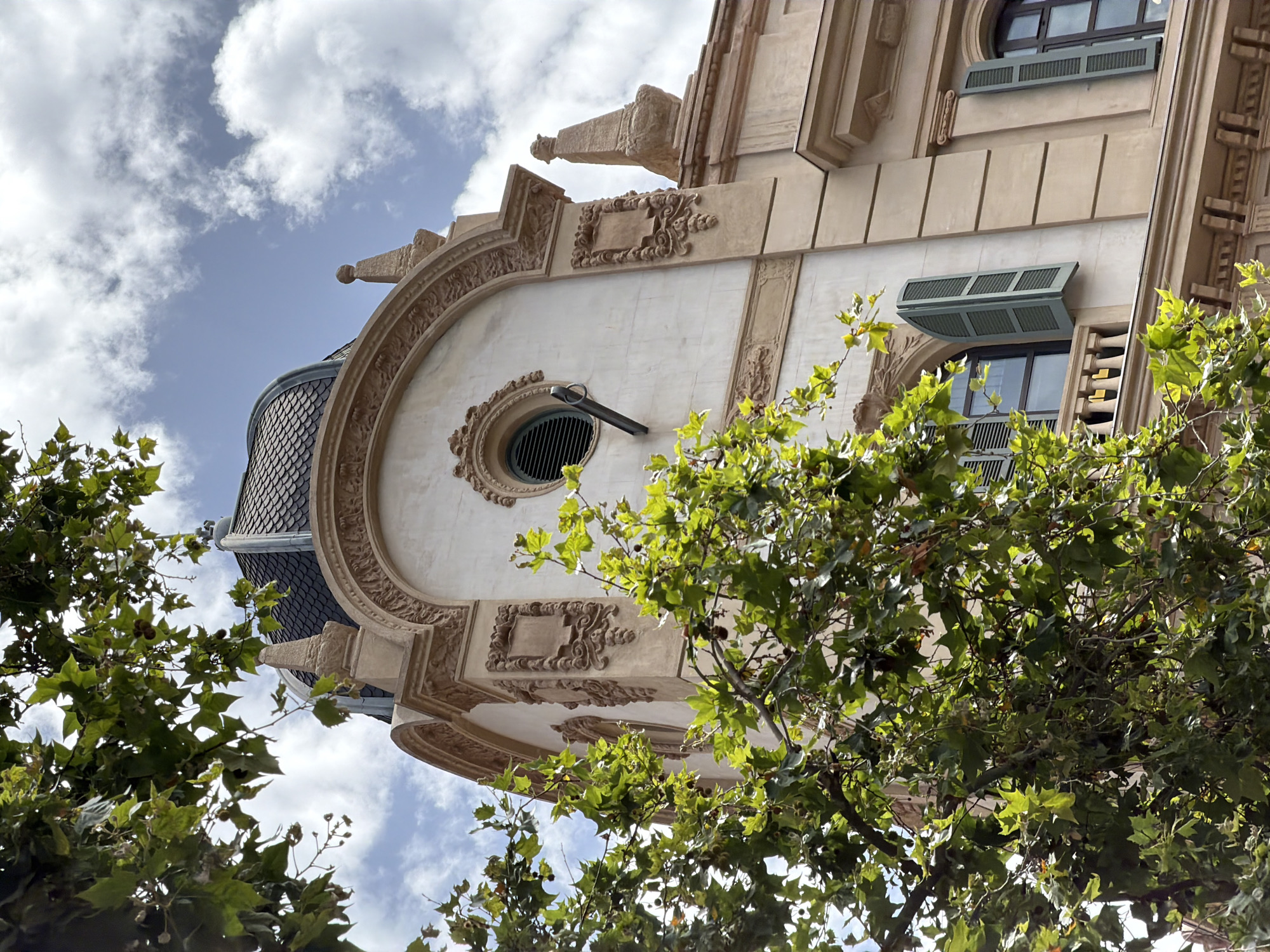 The image captures a close-up view of a section of a building's facade, characterized by ornate architectural details and a dark, tiled dome. The building is rendered in a light beige stone and features intricate carvings, decorative moldings, and a circular window with louvers. A dark metal pole extends from the circular window. Two windows with green shutters are visible on the lower level, one of which is partially obscured by a balcony railing. A vibrant green tree with broad leaves and seed pods is in the foreground, partially obscuring the building. The sky is a light blue with scattered white clouds. This architectural style and dome suggest the building is located in Barcelona, Spain, likely in an area with Beaux-Arts or Art Nouveau influences.