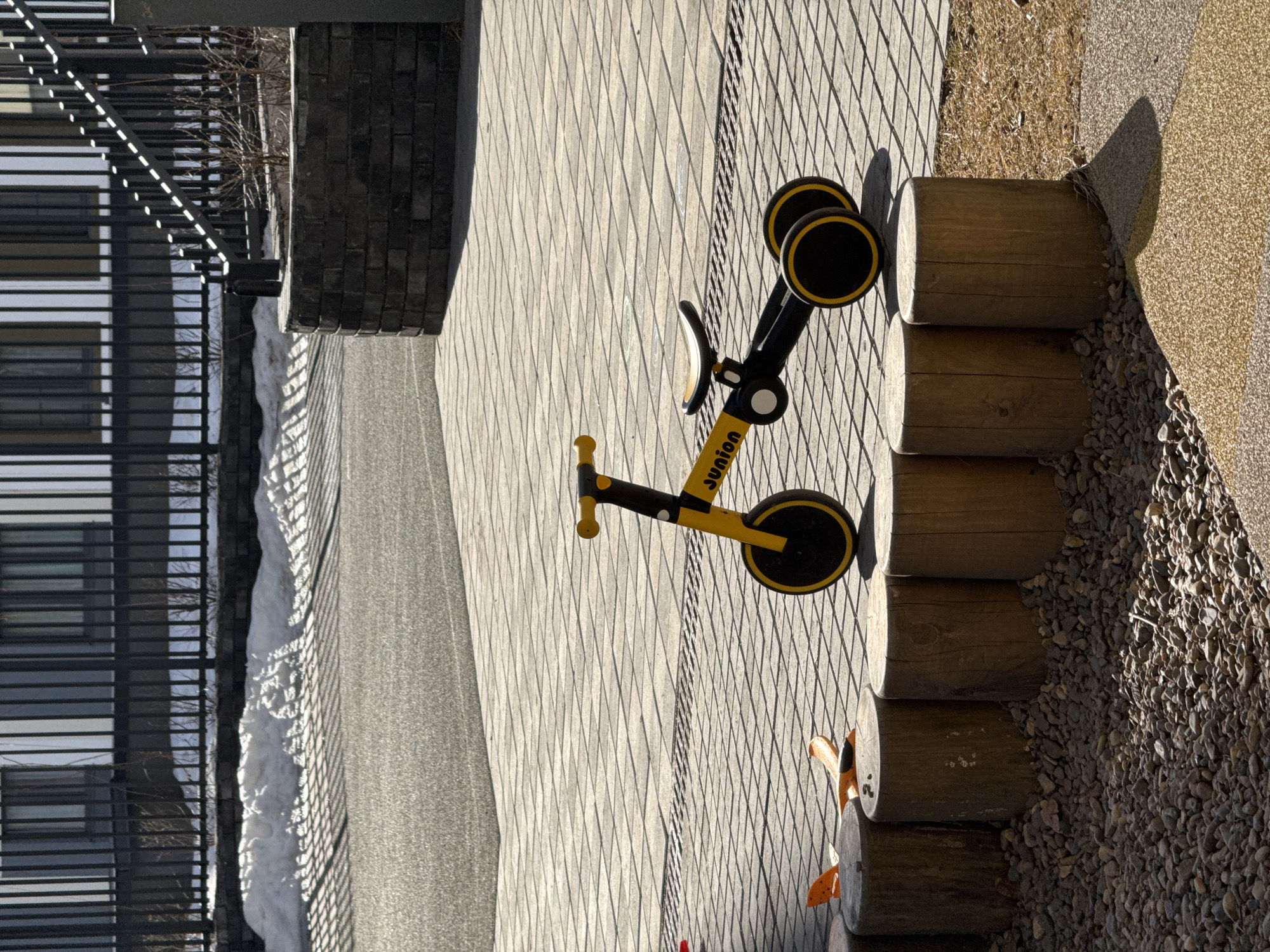A yellow and black balance bike is parked on a brick pathway in an outdoor environment. The bike is oriented diagonally in the frame, with its front wheel near a set of wooden logs stacked to form a low wall. To the right of the logs, there is a gravel area. The pathway is made of gray bricks laid in a repeating pattern. In the background on the left, there is a building with a black metal railing and windows, with a patch of snow visible near the railing. It is daytime, and the scene is well-lit. There are no people visible in the image. The city and country are provided only for context; the visible elements are described regardless.