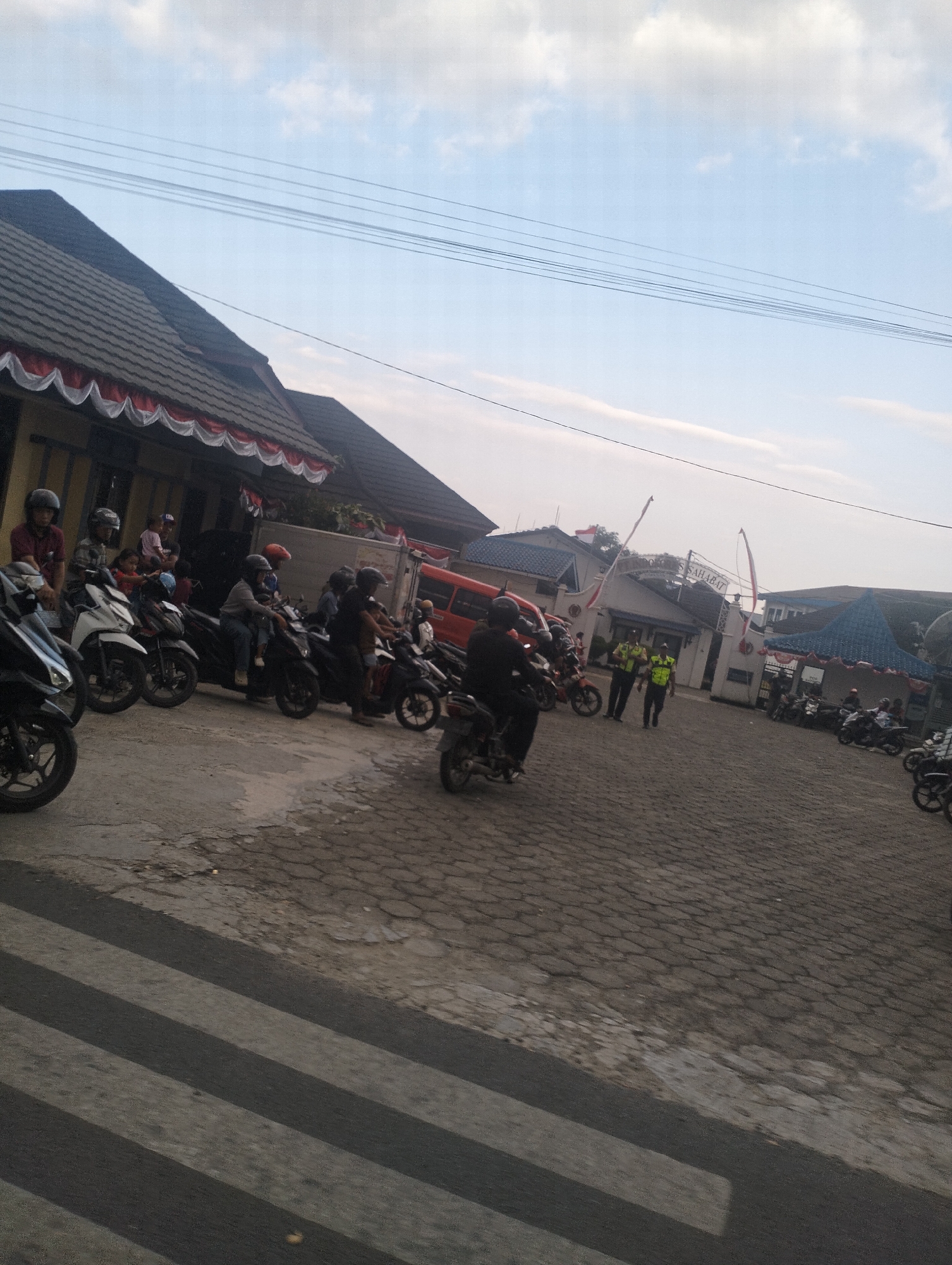 The image captures a scene at what appears to be the entrance to a building complex in Kandang Gampang, Indonesia. Several motorcycles are parked and some are being ridden in a paved outdoor area. In the foreground, a pedestrian crossing is visible on the asphalt. A building with a dark tiled roof and decorative white and red trim is situated on the left side of the frame. To the right, a white gate structure with Indonesian flags and signage is present, indicating an entrance. Two individuals wearing high-visibility vests, possibly security personnel, are standing near the gate. A red truck is also visible parked in the background. The sky is bright with scattered clouds, suggesting daytime.