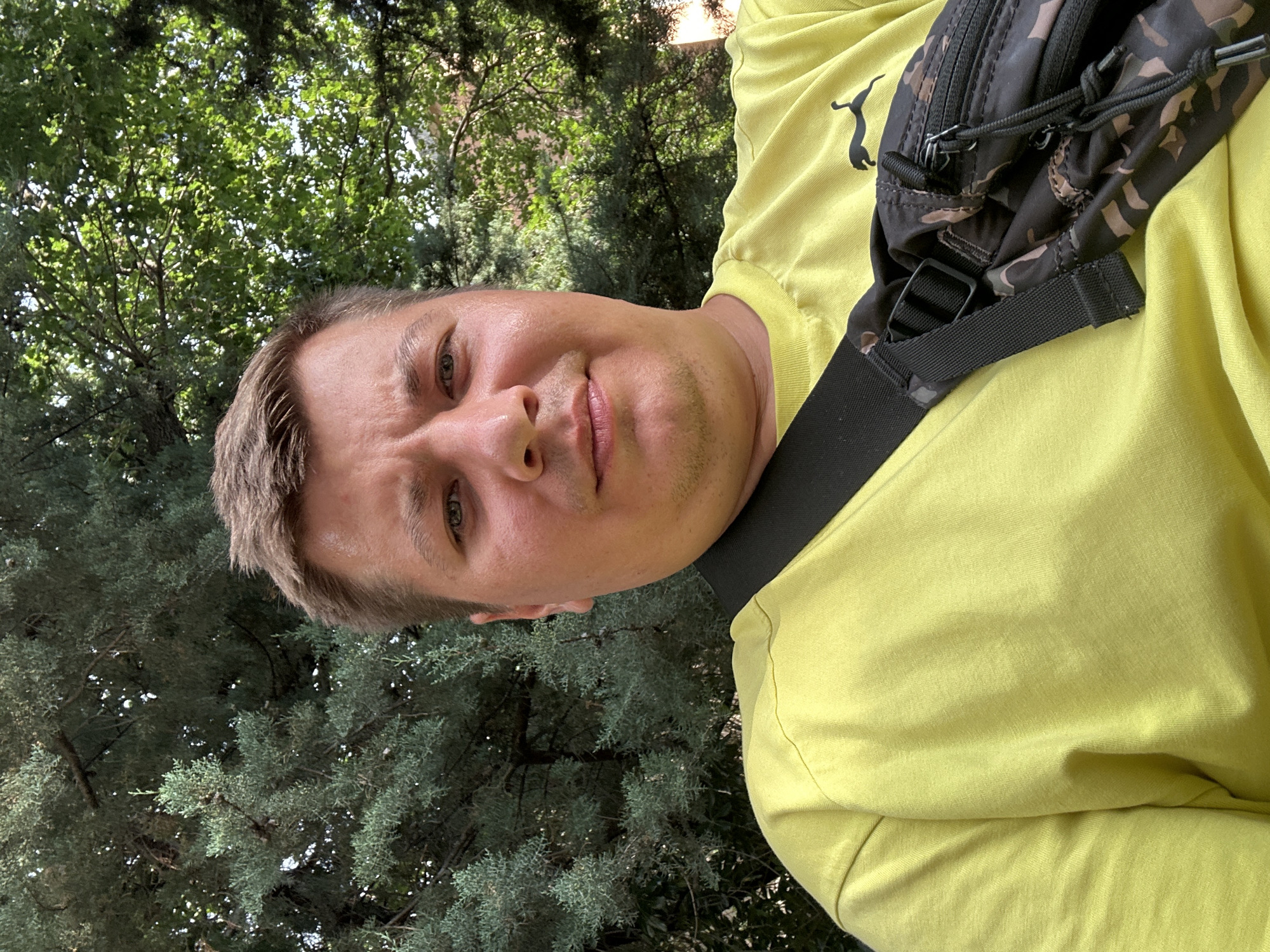 A man is taking a selfie outdoors, with trees in the background. He is wearing a bright yellow t-shirt and a black fanny pack with a camouflage pattern is visible across his chest. The man's face is the primary focus of the image, and he appears to be smiling slightly. The setting suggests it is daytime, likely with pleasant weather, given the outdoor context and the man's attire. The foliage indicates a natural or park-like environment. The image is taken from a low angle, looking upwards towards the man and the trees. There is no visible text in the image. The overall mood is casual and relaxed.