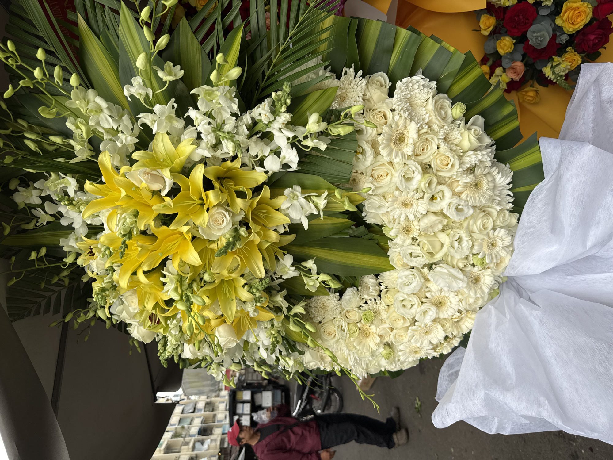 An outdoor flower stall in Nha Trang, Vietnam, displays a large, elaborate funeral wreath composed of predominantly white and yellow flowers, including lilies and roses. The wreath is adorned with lush green foliage. A man in a red cap and maroon jacket is visible in the background, appearing to be holding a plastic bottle. The scene suggests a bustling urban environment with buildings and a motorcycle present. The composition focuses on the floral arrangement, with a white fabric draped across the lower portion of the wreath.