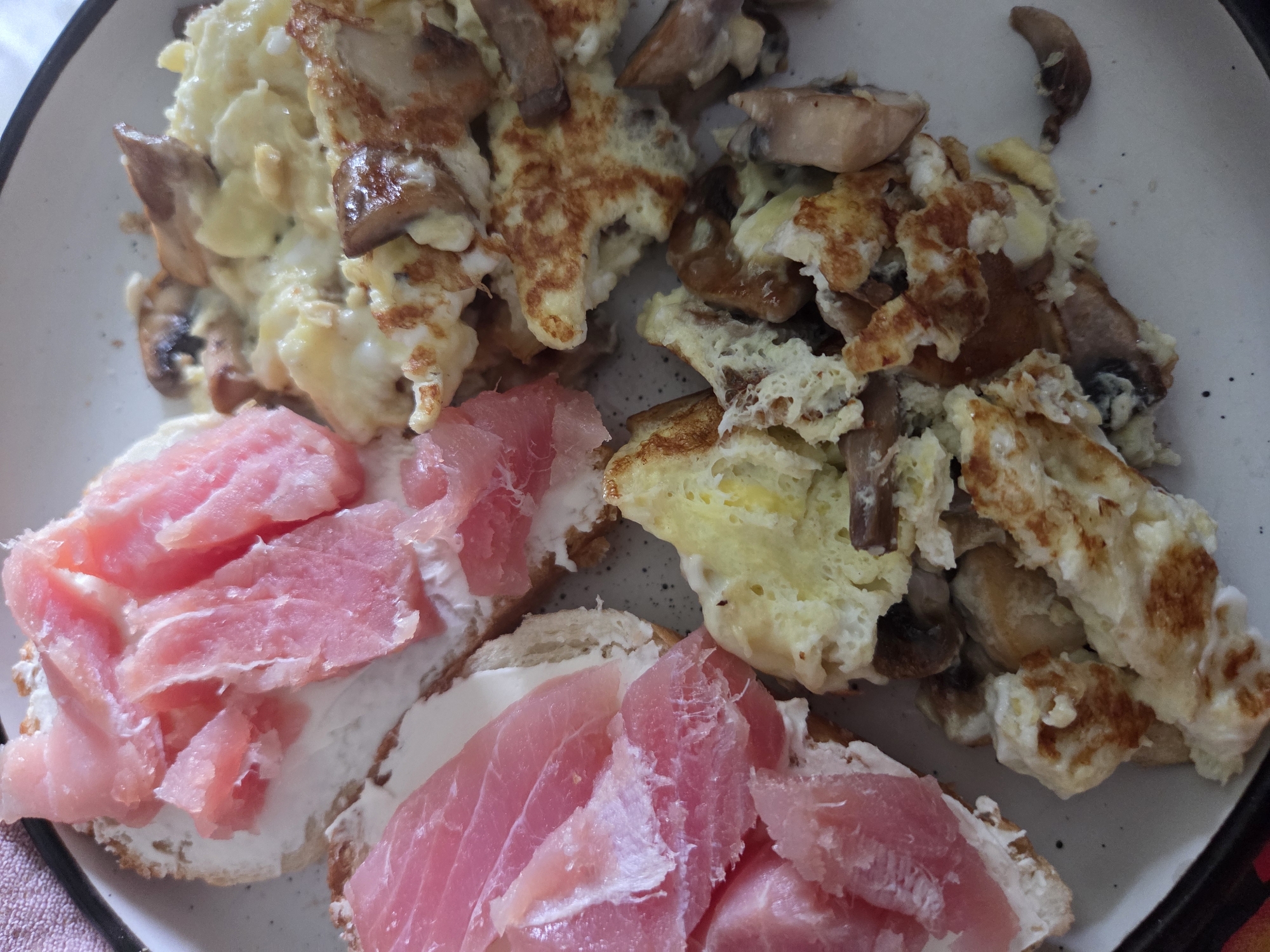 A plate contains scrambled eggs with mushrooms, along with open-faced sandwiches topped with a white spread and pink meat slices. The location is unknown.