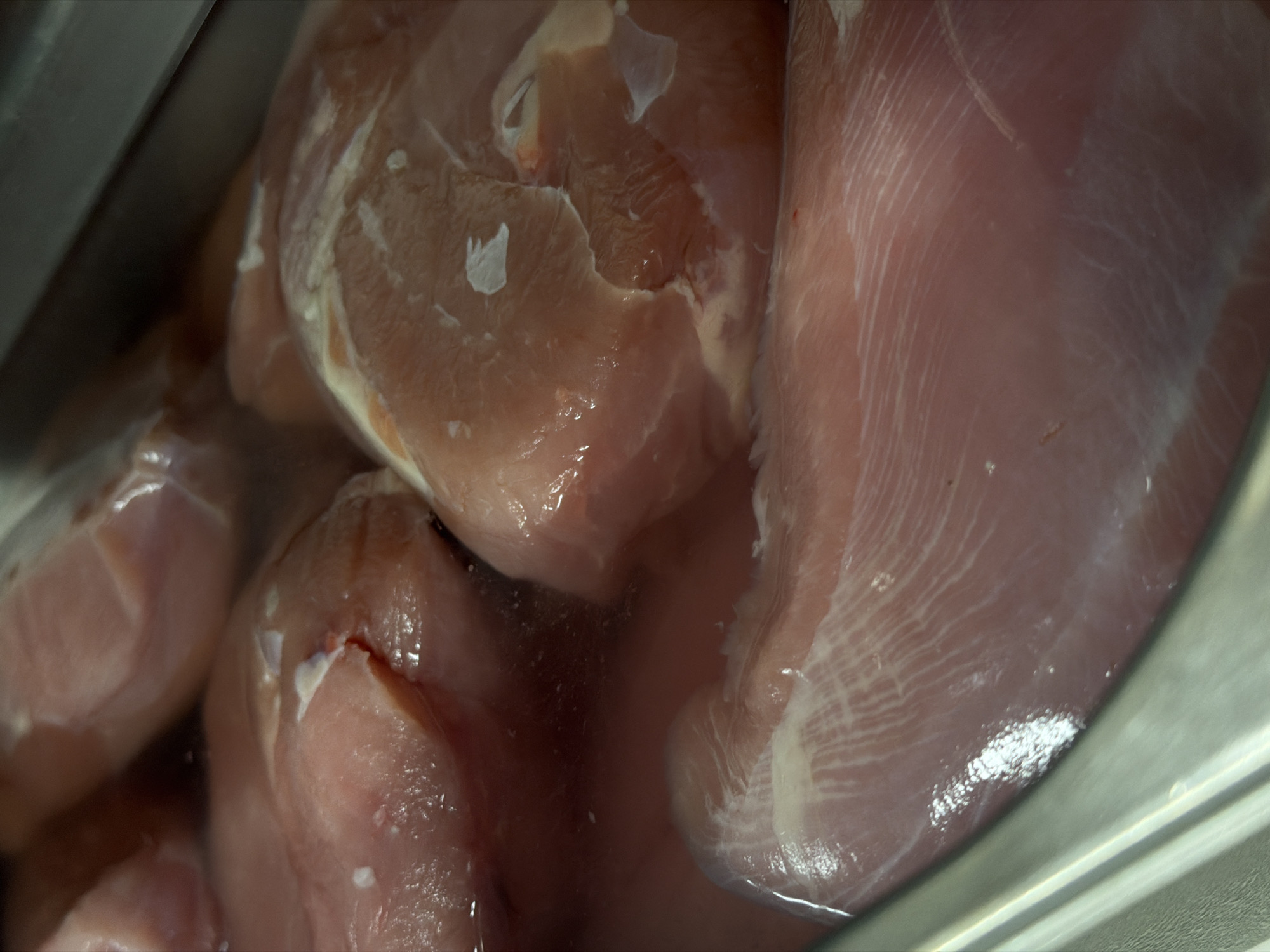 The image depicts several pieces of raw chicken breasts submerged in liquid, likely water. The chicken is arranged in a stainless steel container, possibly a sink. No people are visible. The setting is indoors. The image does not contain sufficient information to determine the specific location in Belgrade, Serbia.