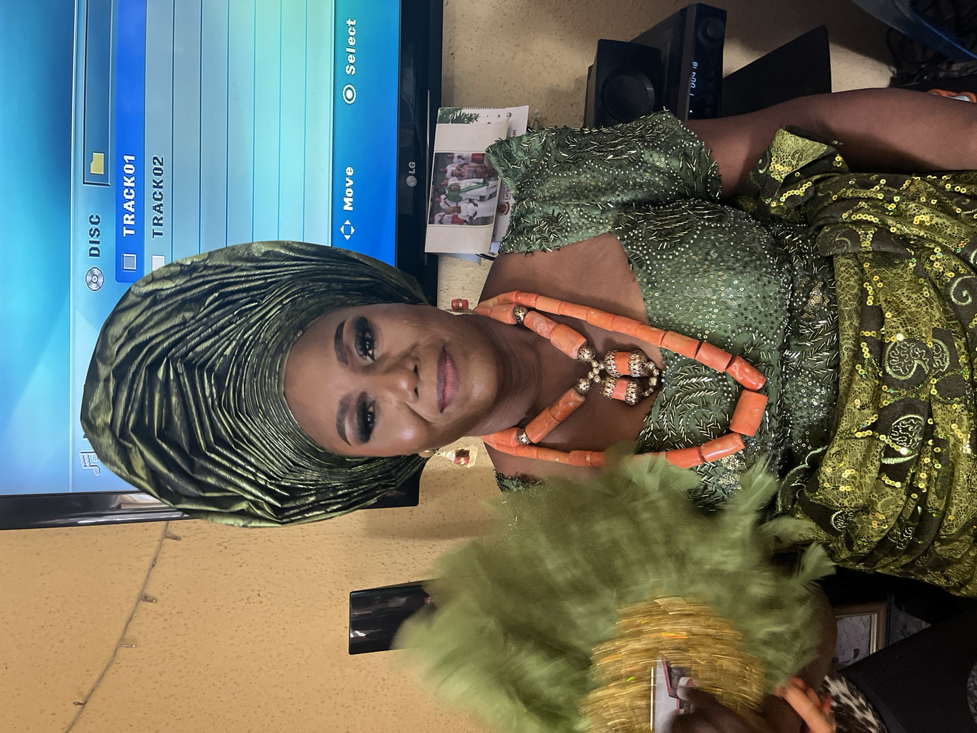 A medium shot captures a woman adorned in traditional Nigerian attire indoors. She wears a green headwrap and a matching embellished dress, complemented by an orange coral necklace. She holds a light green feathery fan. Behind her is a television displaying menu options, and a black speaker. The background appears to be a residence; the specific address or location in Warri, Nigeria, cannot be ascertained from the image.