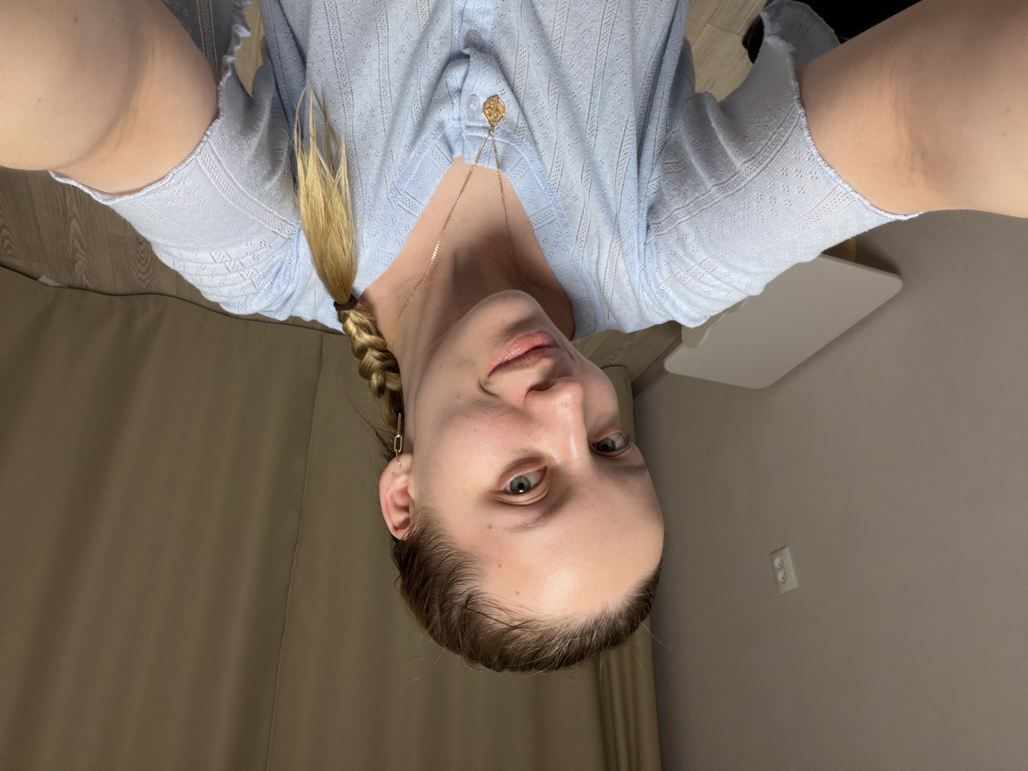 A woman with fair skin and blue-gray eyes is positioned in the center of the image, facing the camera with a slight tilt of her head. Her blonde hair is partially braided, with a loose strand falling beside her face. She wears a light blue button-down shirt with a textured pattern and a delicate gold necklace with a pendant.
The environment consists of a neutral-toned wall in the background and a glimpse of a white shelf-like object. A light switch is visible on the wall. The image is taken from a downward angle.
There is no evidence to indicate the location is Reshetnikovo, Russia.