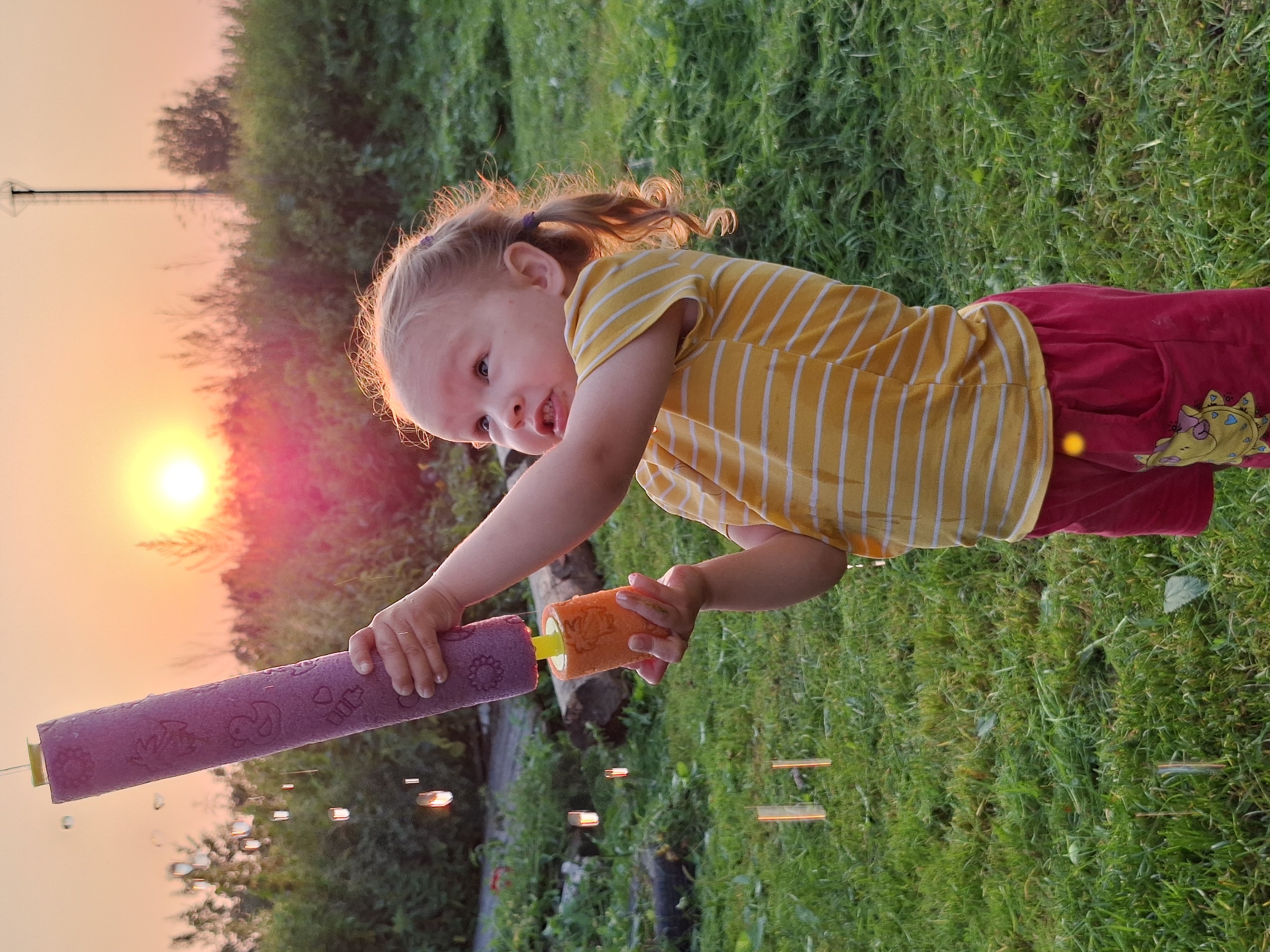 A young child with blonde pigtails is holding a purple toy water gun with an orange handle, aiming it upwards. The child is wearing a yellow and white striped t-shirt and red shorts with a yellow cartoon character design. Water droplets are falling from the water gun and in the background. The child is standing in a grassy area with trees and bushes. The sun is setting in the background, casting a warm glow over the scene. A tall, thin tower-like structure is visible in the distance. The location is described as Unknown, Unknown.