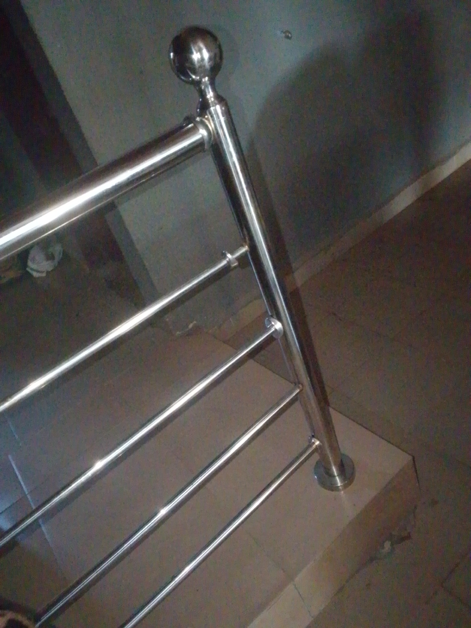 The image shows a close-up of a stainless steel handrail, likely part of a staircase or balcony. The handrail is polished and reflects light, highlighting its smooth surface. It consists of a top horizontal bar and several parallel vertical bars underneath, all connected to a central vertical support post. At the top of the post, there is a decorative spherical cap. The handrail is mounted on a beige tiled surface, which appears to be the edge of a step or platform. In the background, the wall is dark and textured, with some lighter shaded areas suggesting a textured wall or possibly a shadow. The floor in the lower part of the image is also a dark, textured surface, possibly tiles or concrete. There are no people visible in the image. The lighting is artificial, and the scene appears to be indoors, likely in a building. The overall impression is one of modern, clean design. Given the location context of Jalingo, Nigeria, this could be a feature in a residential building, commercial space, or public area. There is no visible text in the image.