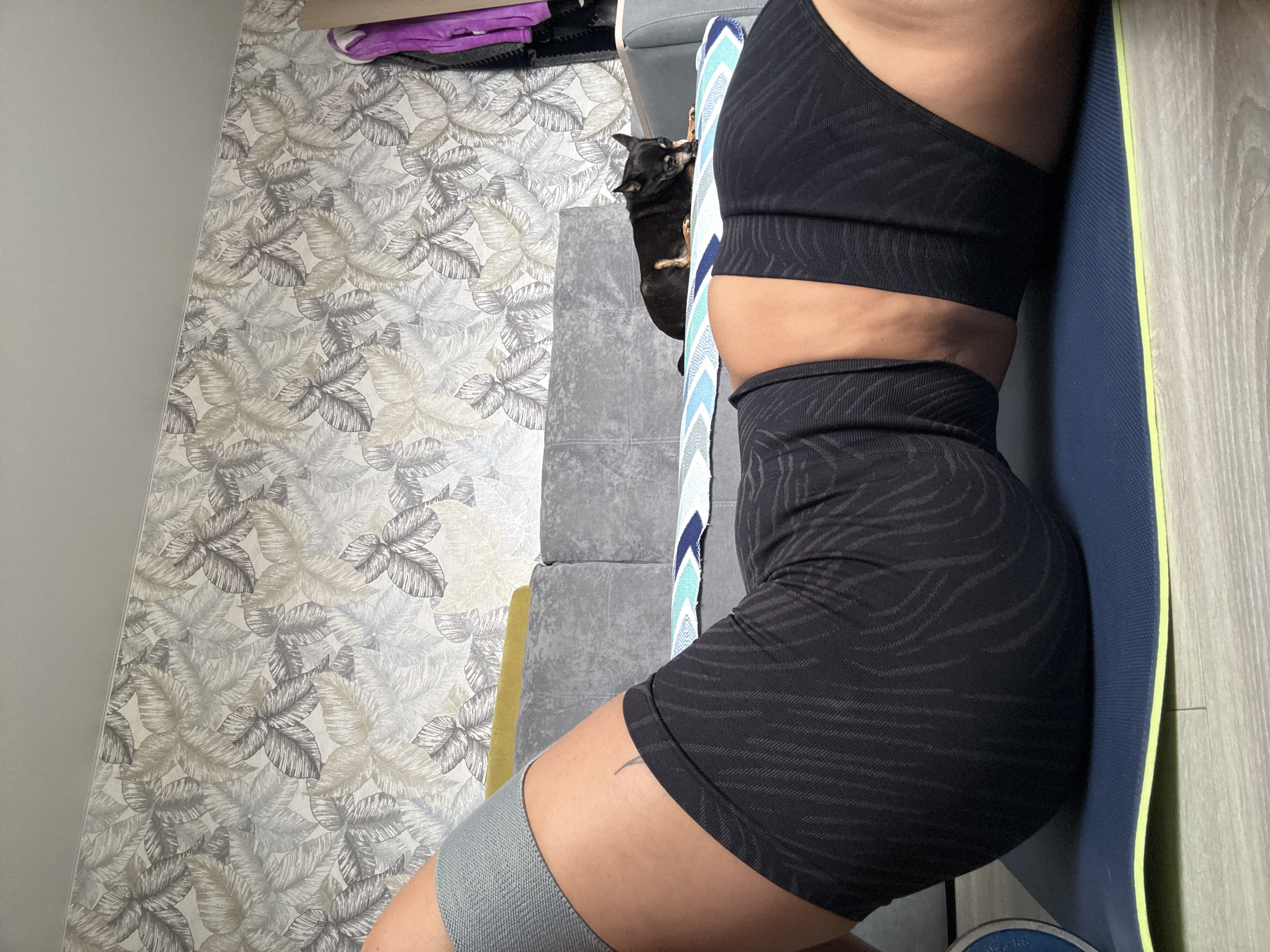 A person is seen in the frame, wearing a black zebra-striped sports bra and matching shorts, along with a gray resistance band on their thigh. They are positioned next to a rolled-up blue and yellow yoga mat, leaning against it. To the left, there is a gray sofa with a small black dog visible on it. The background consists of a wall covered with a leaf-patterned wallpaper. The floor is made of light wood-look panels. The image provides no information to ascertain its location in Kauchuk Stantsiya, Russia.