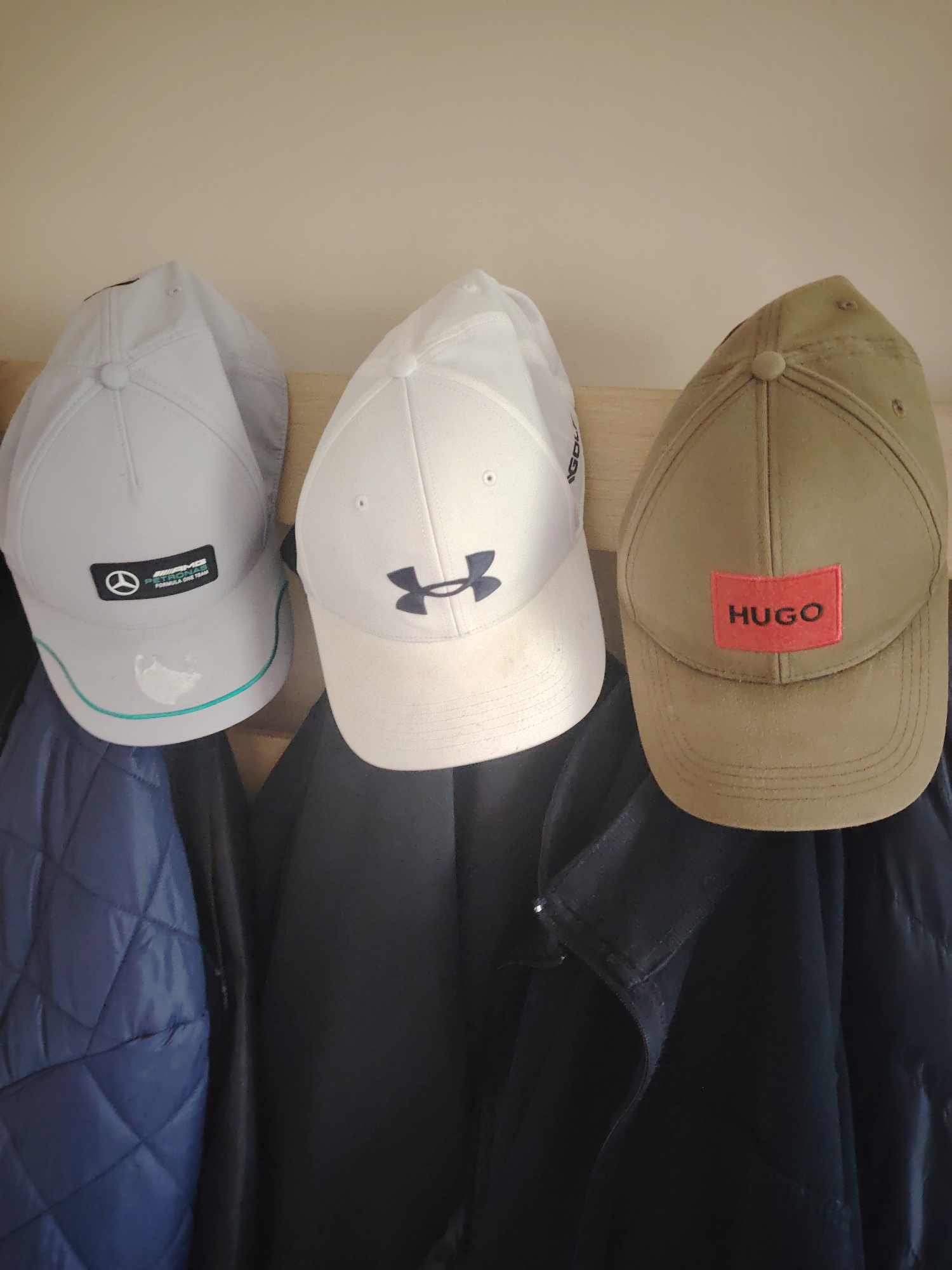 Three baseball caps and three jackets are suspended from an unseen support structure against a plain, light beige wall. The scene is located in Huntingdonshire, United Kingdom.
From left to right:
The leftmost cap is light grey, featuring a black rectangular patch with a white Mercedes-Benz star logo, "AMG PETRONAS", and "FORMULA ONE TEAM" in white text. A thin teal stripe runs along the edge of the brim. Beneath this cap hangs a dark blue quilted jacket.
The central cap is white, displaying a dark blue Under Armour logo. The brim of this cap shows minor discoloration. Beneath this cap hangs a dark, unidentifiable jacket or coat.
The rightmost cap is khaki-colored, featuring a prominent red rectangular patch with the word "HUGO" in black capital letters. The surface of this cap appears slightly dusty or worn. Beneath this cap hangs another dark, unidentifiable jacket or coat, with a collar detail visible on the right side.
The wall behind the items is a consistent light beige tone. No individuals are present in the image.