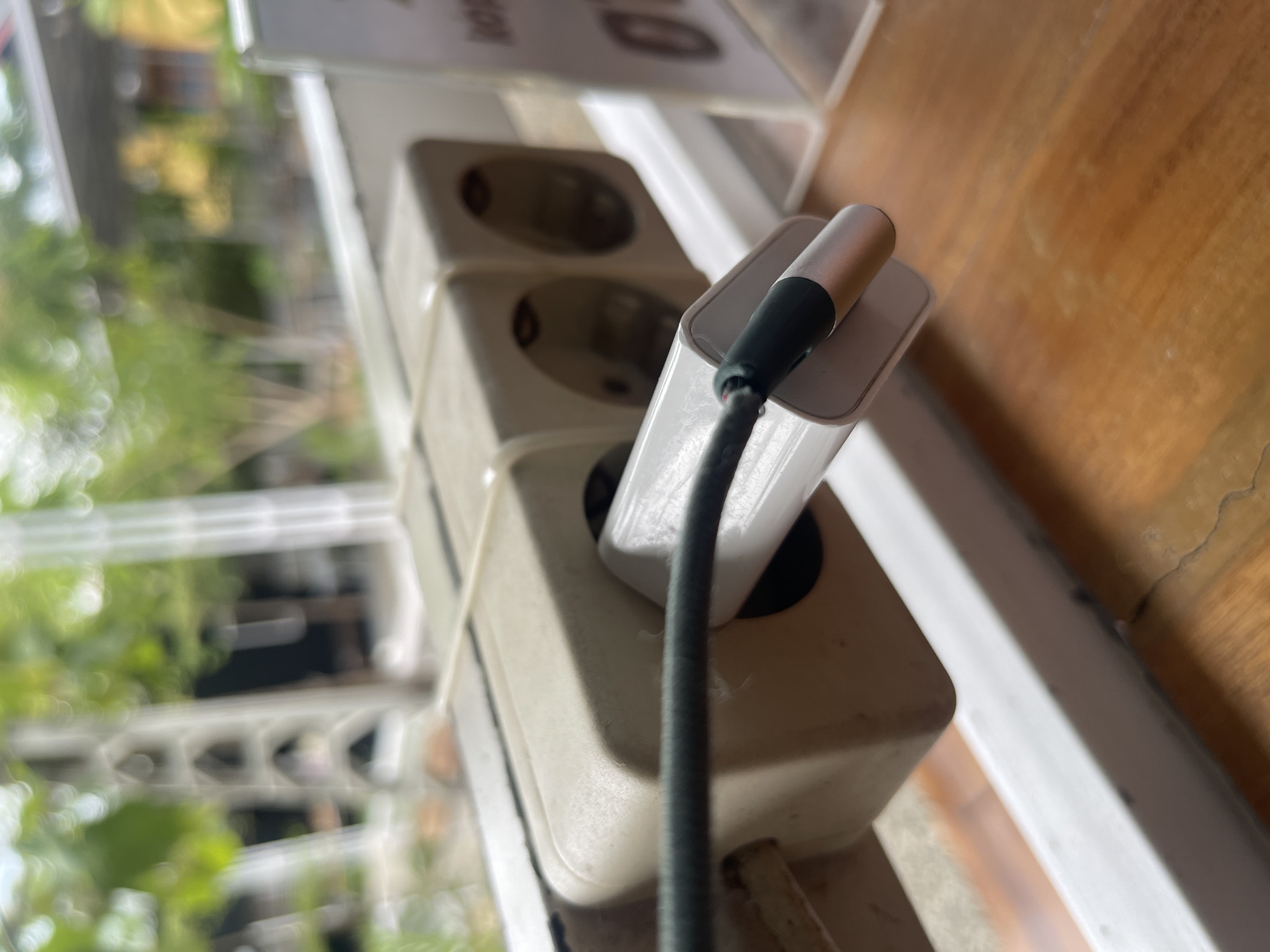 In Pulokiton, Indonesia, a white electrical extension cord with two outlets is secured with two white zip ties to a white horizontal bar. A white phone charger is plugged into the leftmost outlet, with a dark green cable extending from it. The background is blurred, showing greenery and a building, suggesting an outdoor or semi-outdoor setting. The image is taken from a slightly elevated angle, focusing on the electrical outlet and charger.