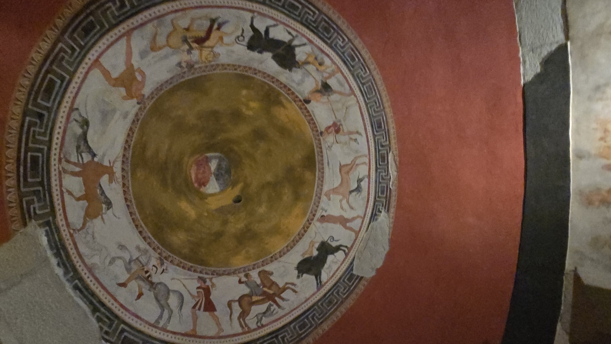 This is a photograph of the ceiling fresco in the Tomb of the Griffins, located in the Ancient City of Augusta, in modern-day Svilengrad, Bulgaria, not Konstantinovo. The circular fresco depicts a mythological scene with human figures and animals. In the center, there is a circular area with a reddish-brown hue, bordered by a lighter colored circular band. Radiating outward from the center, the fresco is divided into concentric rings filled with imagery. The outermost ring features a decorative border with geometric patterns and a band of red. Within this border, there are scenes of what appears to be a hunt or battle, with figures of humans, some on horseback, engaged with animals such as horses, dogs, and a boar. The figures are rendered in a classical style with distinct outlines and muted colors. The background of these scenes is a light, almost off-white color. The overall impression is of a dynamic and elaborate ancient artwork adorning the ceiling of a burial chamber.