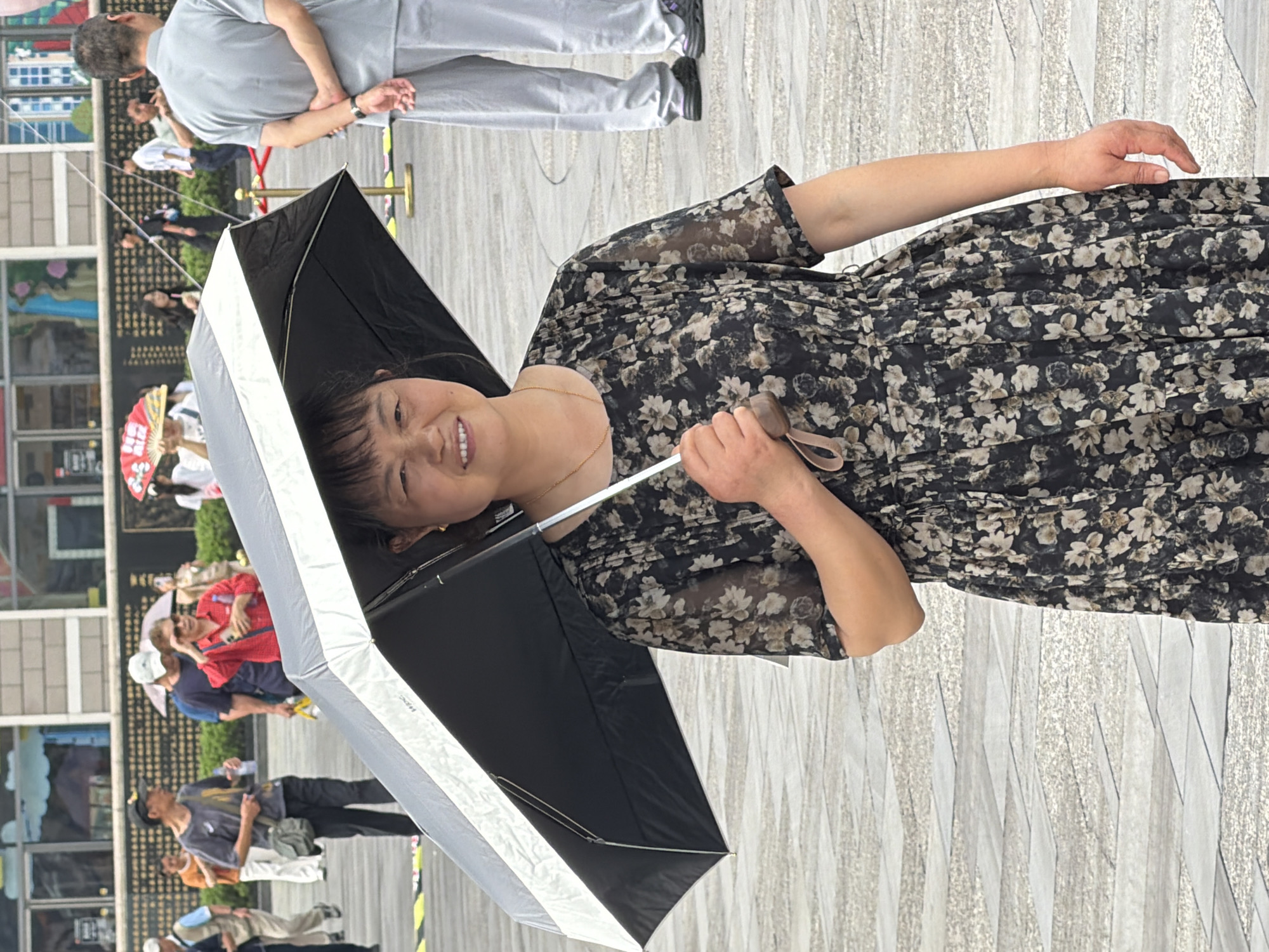 In Yuzhong District, China, a woman holding a black and white umbrella stands in the foreground, facing the camera with a smile. She is wearing a dark, floral-patterned dress. Behind her, the scene unfolds with various individuals in the background, some holding umbrellas and parasols. The environment appears to be an outdoor public space with paved ground.