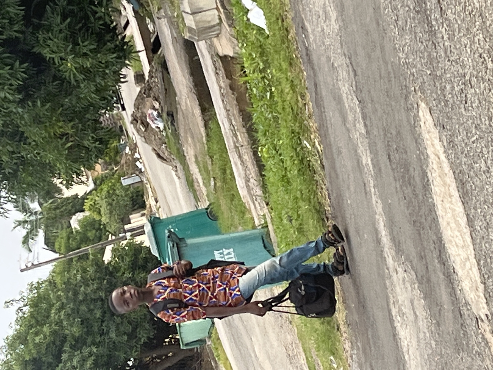 A young boy walks on a paved road in Abuja, Nigeria. He is wearing a patterned shirt, blue jeans, and sandals, and carrying a backpack and a black duffel bag. Two green trash bins are visible behind him. The environment appears to be an urban setting with overgrown grass, trees, and debris on the side of the road. The sky is overcast.