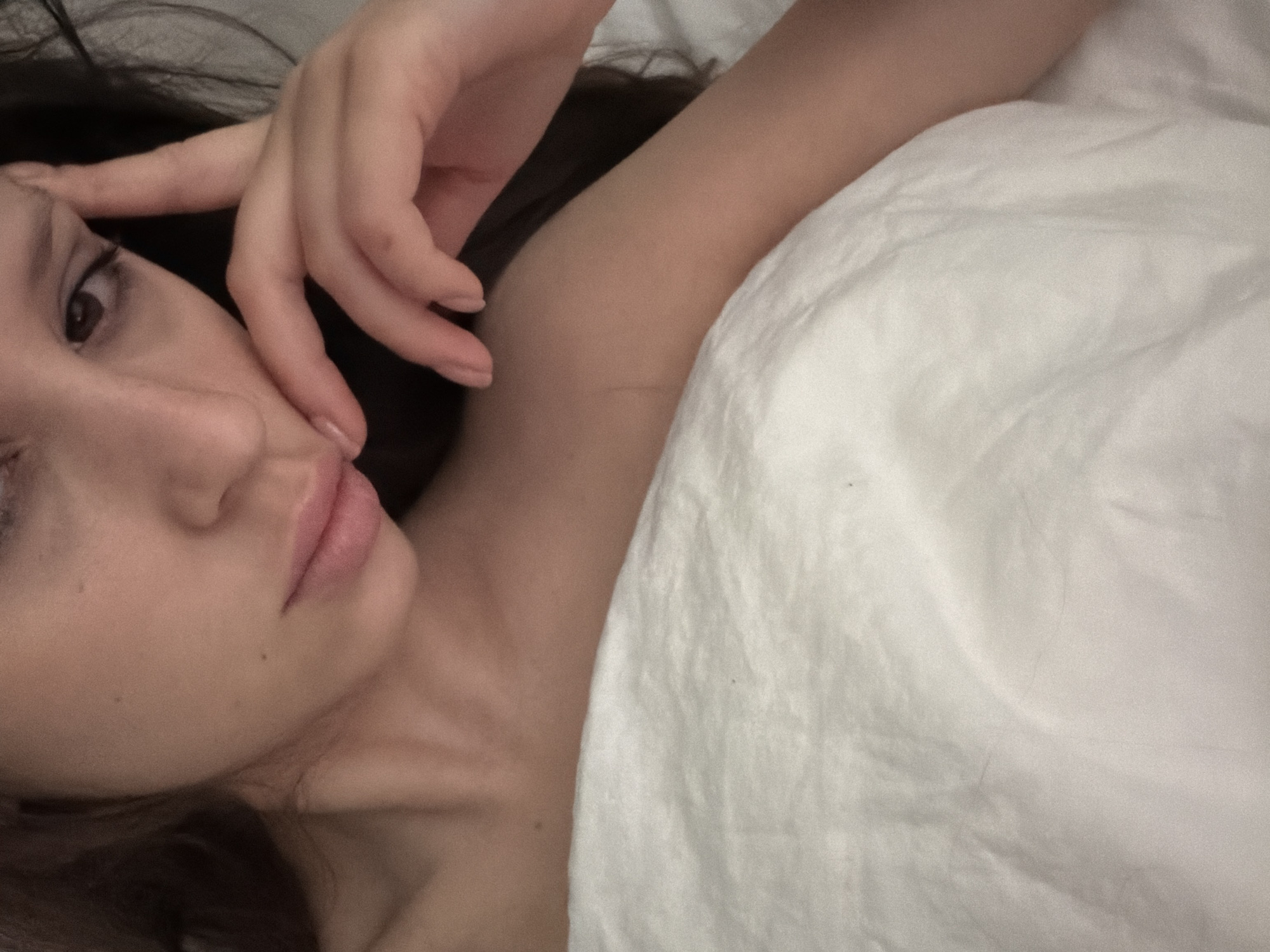 The image shows a close-up, overhead view of a young person lying in bed. Their head is tilted, and their right hand is raised to their forehead, with fingers touching their temple. Their eyes are partially closed, and their lips are slightly parted, suggesting a pensive or tired expression. The person has fair skin and dark hair. They are covered by a white sheet, which is rumpled around them. The lighting is soft and diffused, creating a calm and intimate atmosphere. The background is dark and out of focus. There are no visible elements that indicate the location in Khimki, Russia.