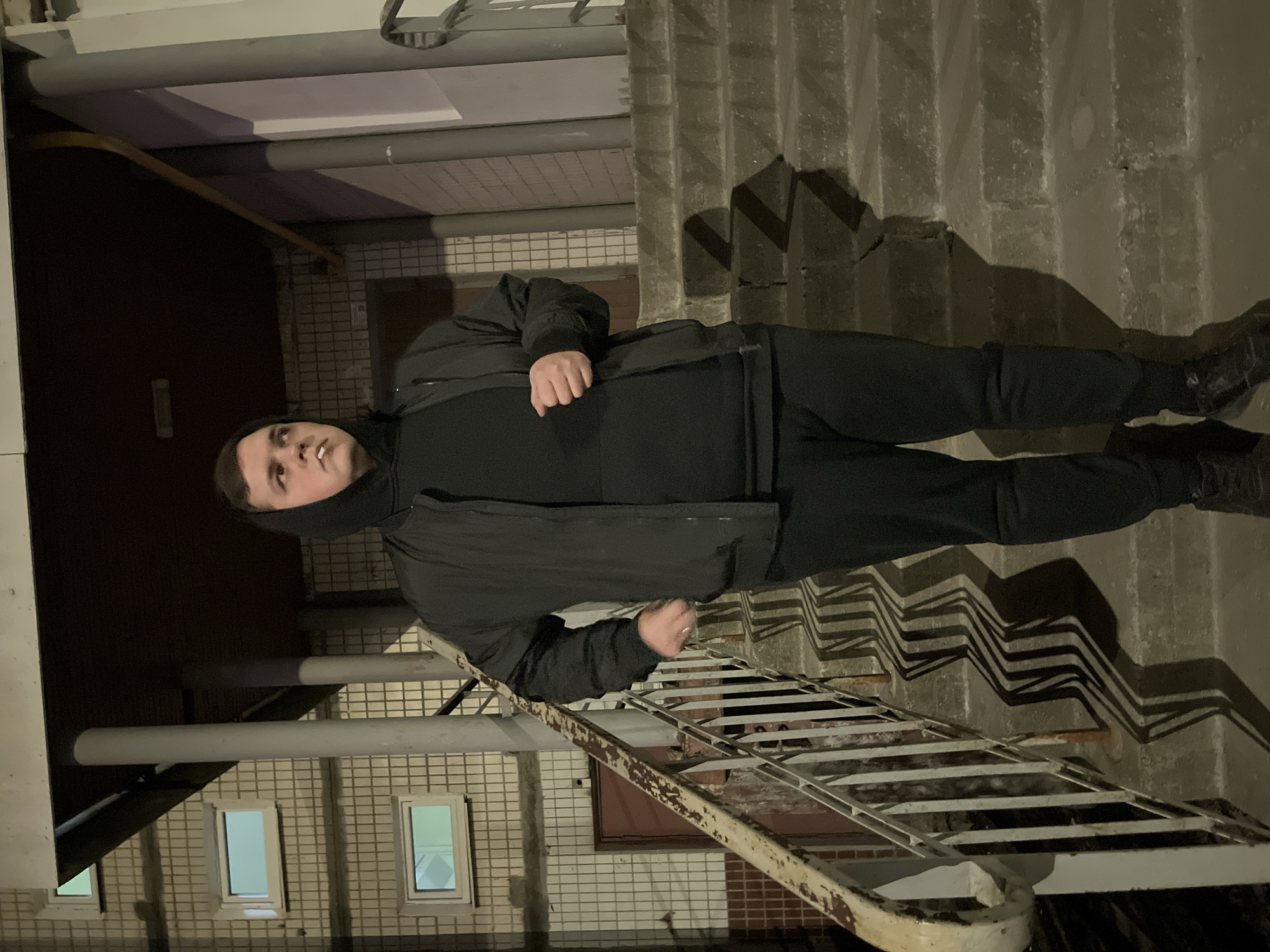 A man stands on an outdoor metal staircase in Klin, Russia. He wears black clothing and shoes. The building behind the staircase has windows and brickwork. The staircase casts shadows on a concrete wall.