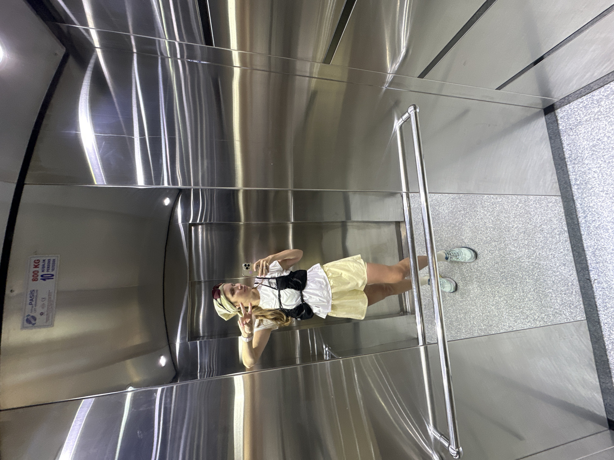 The photograph shows a woman taking a selfie in an elevator, which appears to be located in Fethiye, Turkey, given the signage in the image. The woman is wearing a white T-shirt, yellow shorts, and a headscarf. She has her phone in her hand and is making a peace sign. The elevator walls are stainless steel, and the floor is gray speckled tile. There is a metal grab bar to her right. A sign on the left wall displays the elevator's capacity as 800 kg and 10 persons, along with the word "PASIS."
