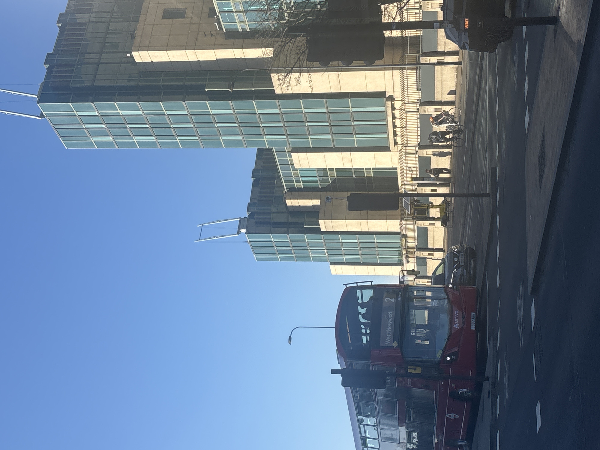 The image shows a street scene in London, United Kingdom. Two modern glass and concrete buildings dominate the left side of the frame, set against a clear blue sky. A red double-decker bus, marked as "Arriva" and displaying "West Norwood", is in the foreground on the right, appearing to be in motion on the road. Other vehicles, including a car, are also visible on the road. Pedestrians and cyclists are present in the background near the buildings. Traffic lights and street signs are visible along the street.