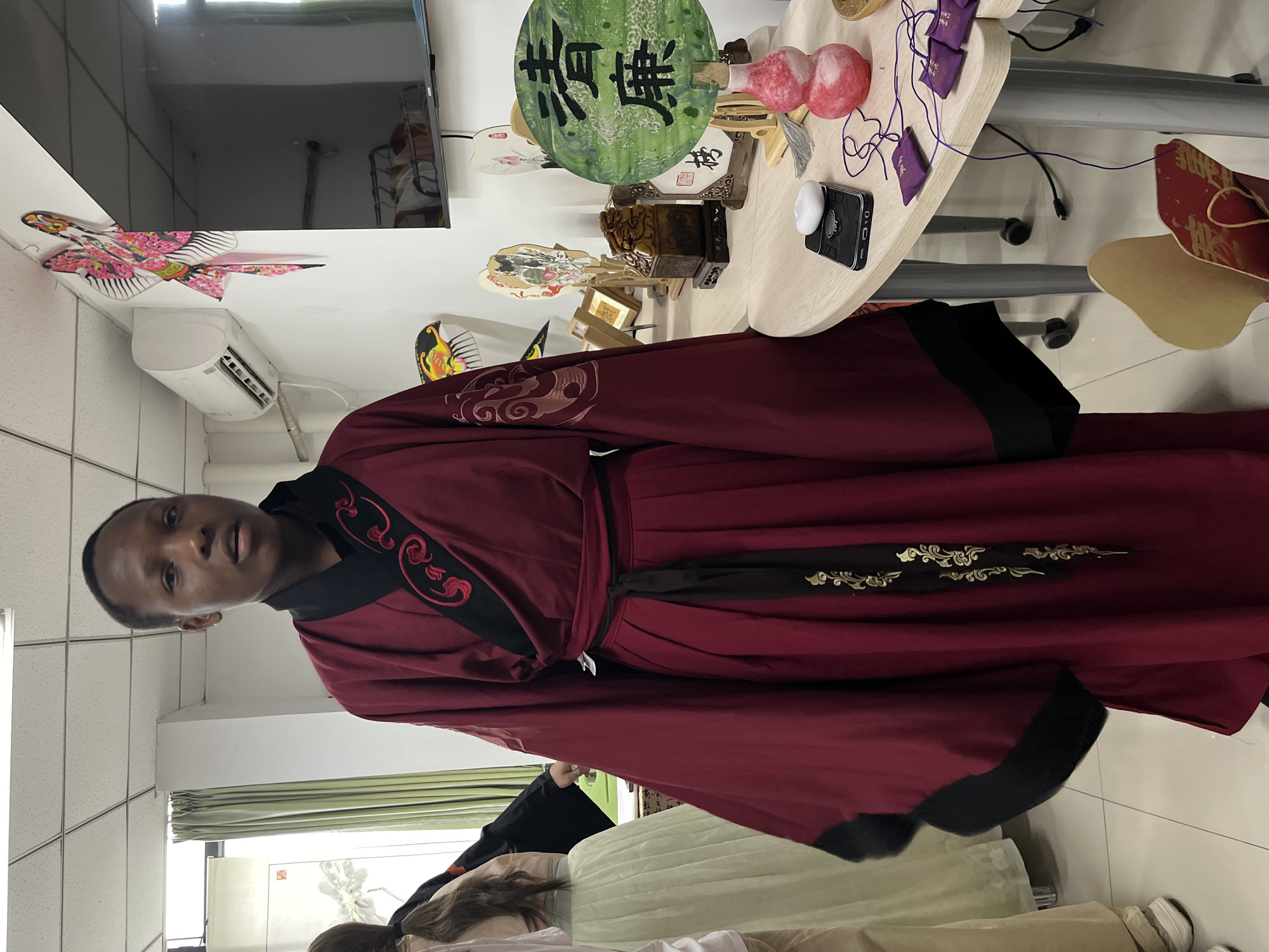 A man wearing a red and black Hanfu stands indoors. The location appears to be a room in Jiangjin, China, based on the presence of Chinese fans, calligraphy, and other cultural artifacts. The man is wearing a maroon robe with black trim and decorative embroidery. There is a table with various items on it, including a green fan with Chinese characters, decorative gourds, and other objects. The room also contains a television, an air conditioner, and other furniture. Other people are partially visible in the lower part of the image.