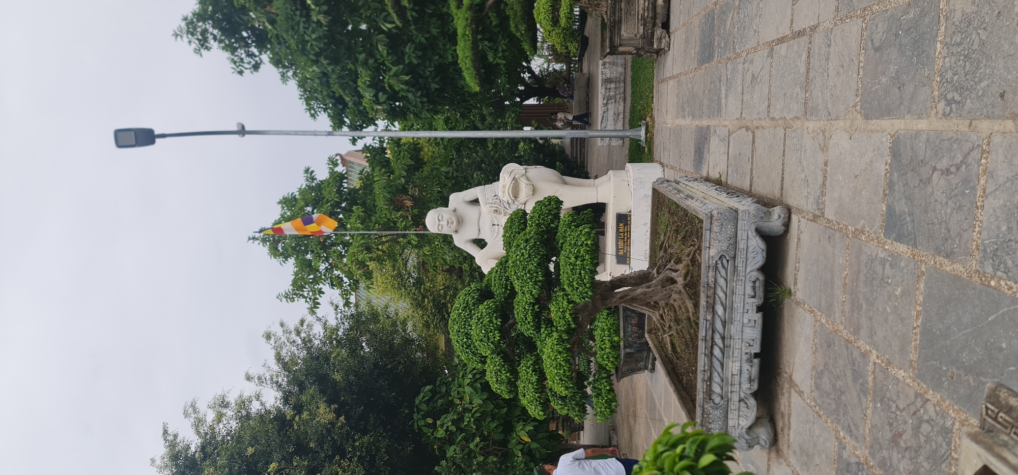 The image displays an outdoor scene in Da Nang, Vietnam, featuring a white statue of a man, likely an Arhat (Buddhist enlightened one), on the right side. The statue, depicted with a calm expression, appears to be carrying a large object, possibly a basket or bowl, on its back or shoulder.
In front of the statue is a decorative, rectangular planter containing a meticulously pruned, bonsai-style tree with dense green foliage and exposed roots. At the base of the statue, a black plaque is visible, bearing text in Vietnamese. The prominent inscription reads "BẢI KIỀU LA HÁN," identifying the figure, followed by "Năm Ất Mùi - 2015," indicating its dedication or creation year as 2015. Smaller text below is partially obscured but appears to be a dedication.
To the left of the statue is a paved pathway made of large, square stone slabs. The background is filled with lush green trees and diverse foliage, with glimpses of distant buildings through the canopy. A tall, grey street lamp pole stands prominently in the mid-ground. Further back and to the left of the statue, a colorful Buddhist prayer flag is affixed to a pole, displaying horizontal stripes of yellow, white, orange, blue, and red.
The sky is overcast, suggesting a cloudy day with diffused light. No specific activity or event is taking place; the scene appears tranquil. A person in a white shirt is partially visible in the bottom left corner, and other distant figures can be discerned in the background.